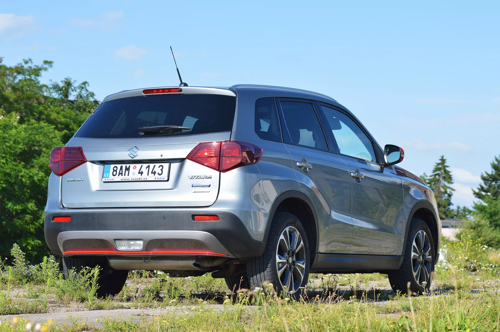 Suzuki Vitara 2018 uchwycona z prawego tylnego kąta trzech czwartych, podkreślająca design tylnych świateł, tylnego okna i profilu pasażerskiej strony. Obraz pokazuje nowoczesne udogodnienia kompaktowego SUV-a, takie jak antena dachowa do funkcji technologicznych, tylne czujniki parkowania i stylowe felgi aluminiowe. Pojazd jest ustawiony na nieutwardzonej powierzchni w wiejskim otoczeniu, co wskazuje na jego zdolności terenowe. Idealne dla kategorii motoryzacyjnych, SUV i obrazów terenowych pojazdów.