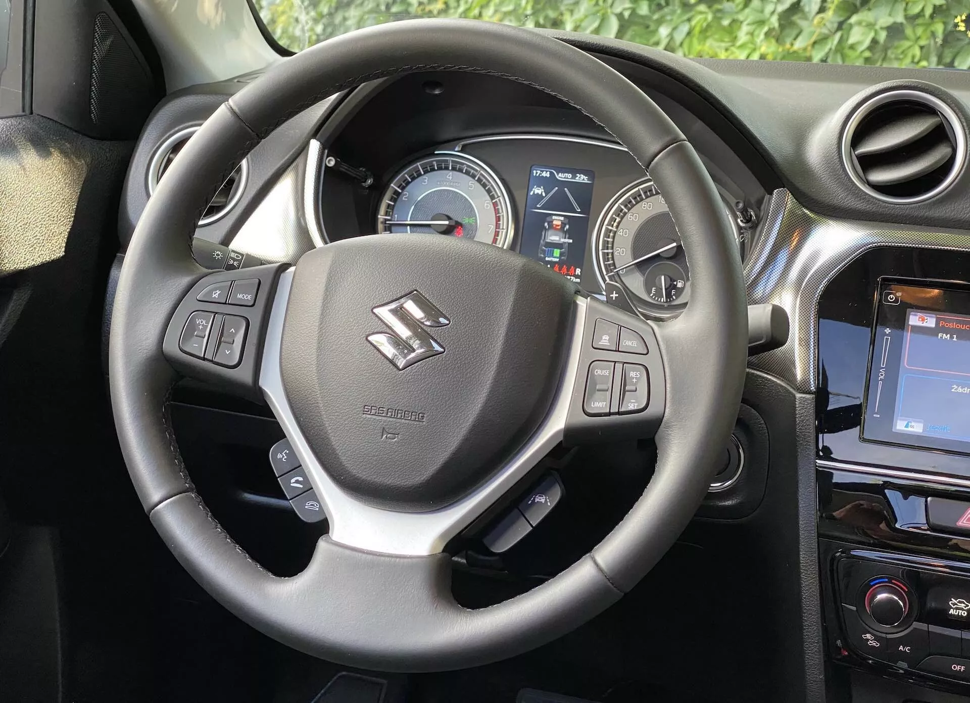Wnętrze Suzuki Vitara 2018, ukazujące perspektywę kierowcy z naciskiem na kierownicę i deskę rozdzielczą. Kierownica, umieszczona w centrum obrazu, posiada podpory na kciuki i przyciski do obsługi audio, głosowych komend oraz tempomatu. Za nią znajduje się zestaw wskaźników z analogowym prędkościomierzem i częściowym wyświetlaczem cyfrowym dostarczającym informacje kierowcy. Po prawej stronie, na środkowej konsoli, widoczny jest system infotainment dotykowy, co wzmacnia nowoczesny wygląd samochodu.