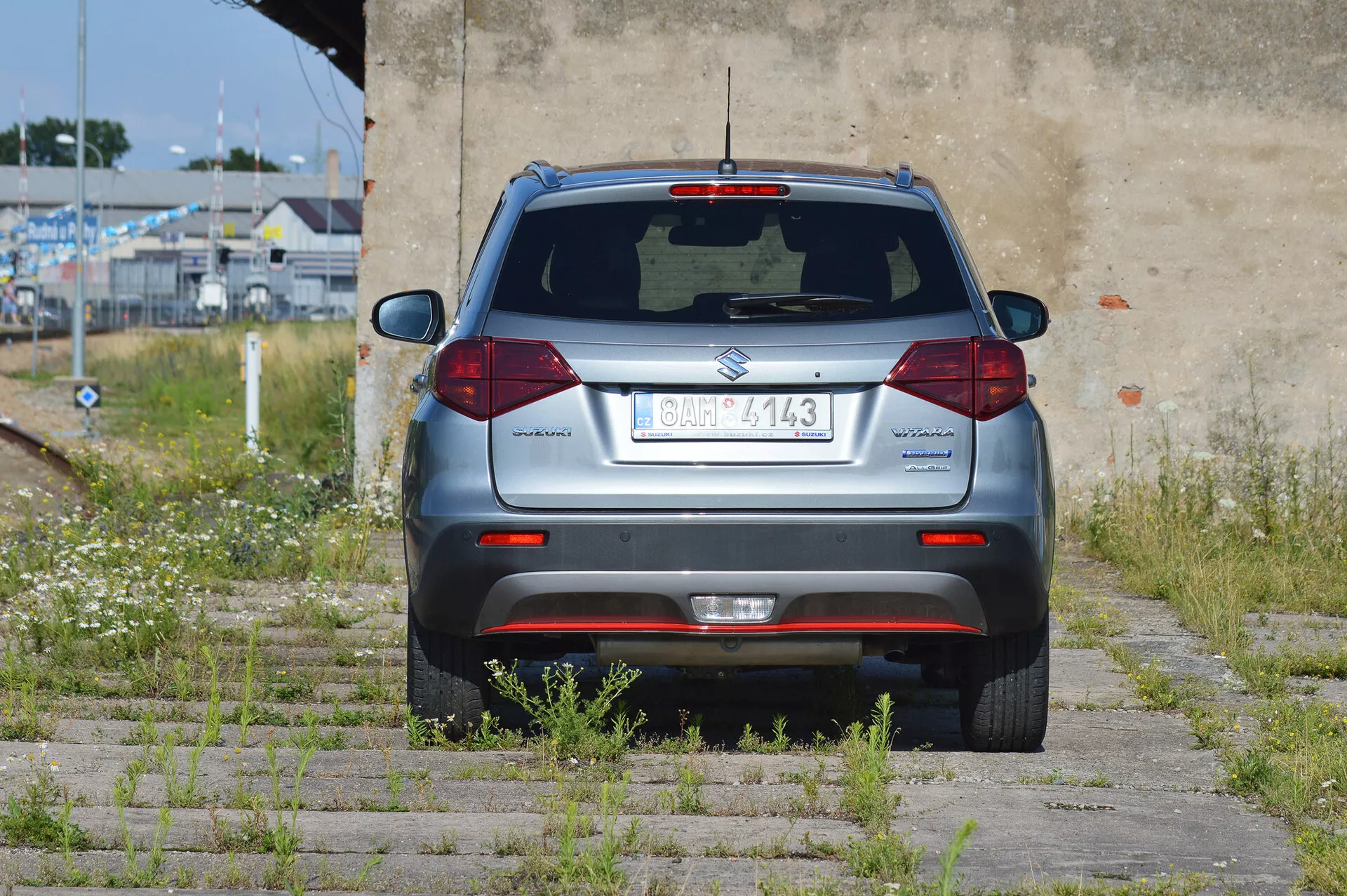 Ujęcie od tyłu pod kątem srebrnego kompaktowego SUV Suzuki Vitara 2018 zaparkowanego na trawie i betonowej kostce. Widoczna jest cała tylna część pojazdu, z centralnie umieszczoną tablicą rejestracyjną, tylną wycieraczką, trzecim światłem stopu, standardowymi halogenowymi światłami tylnymi, odblaskami na zderzaku, czujnikami parkowania i rurą wydechową po lewej stronie. Pojazd znajduje się w półopuszczonym obszarze z przerośniętą roślinnością i zniszczoną ścianą.