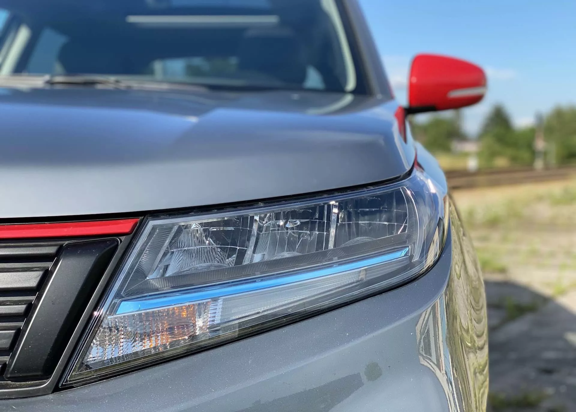 Zbliżenie przedniego lewego narożnika Suzuki Vitara 2018, skupiające się na szczegółach takich jak LED-owe reflektory z światłami dziennymi, częściowo widoczna kratka z pionowymi listwami, delikatnie nachylona maska, metaliczna farba, kontrastujące czerwone lusterko boczne prawdopodobnie z wbudowanymi kierunkowskazami oraz rozmyte tło podkreślające elegancki design i aerodynamiczną wydajność.