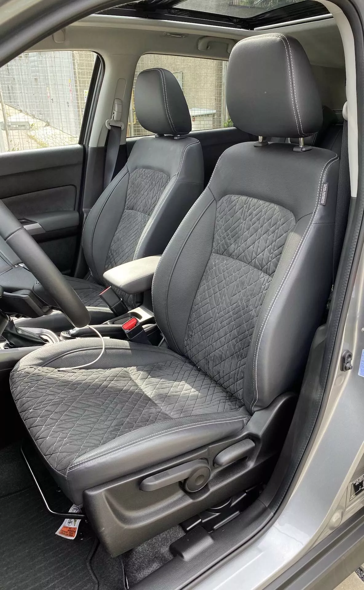 Wnętrze Suzuki Vitara 2018, wyróżniające nowoczesne przednie siedzenia typu kubełkowego z perspektywy nieco powyżej poziomu siedzenia. Fotele są tapicerowane czarną skórą z pikowanym wzorem w romby oraz gładkimi skórzanymi bokami, oferując równowagę między komfortem a sportowością. Czerwona etykieta na siedzeniach sugeruje dodatkowe funkcje, takie jak boczne poduszki powietrzne lub podgrzewane siedzenia. Widoczna jest konsola środkowa z skórzanym podłokietnikiem, a także kabel ładujący na siedzeniu pasażera, co wskazuje na obecność portów ładowania. Klamry pasów bezpieczeństwa dopasowują się do kolorystyki wnętrza w kolorze czarnym. Widać również część deski rozdzielczej oraz słupek B, co sugeruje przestronną kabinę.
