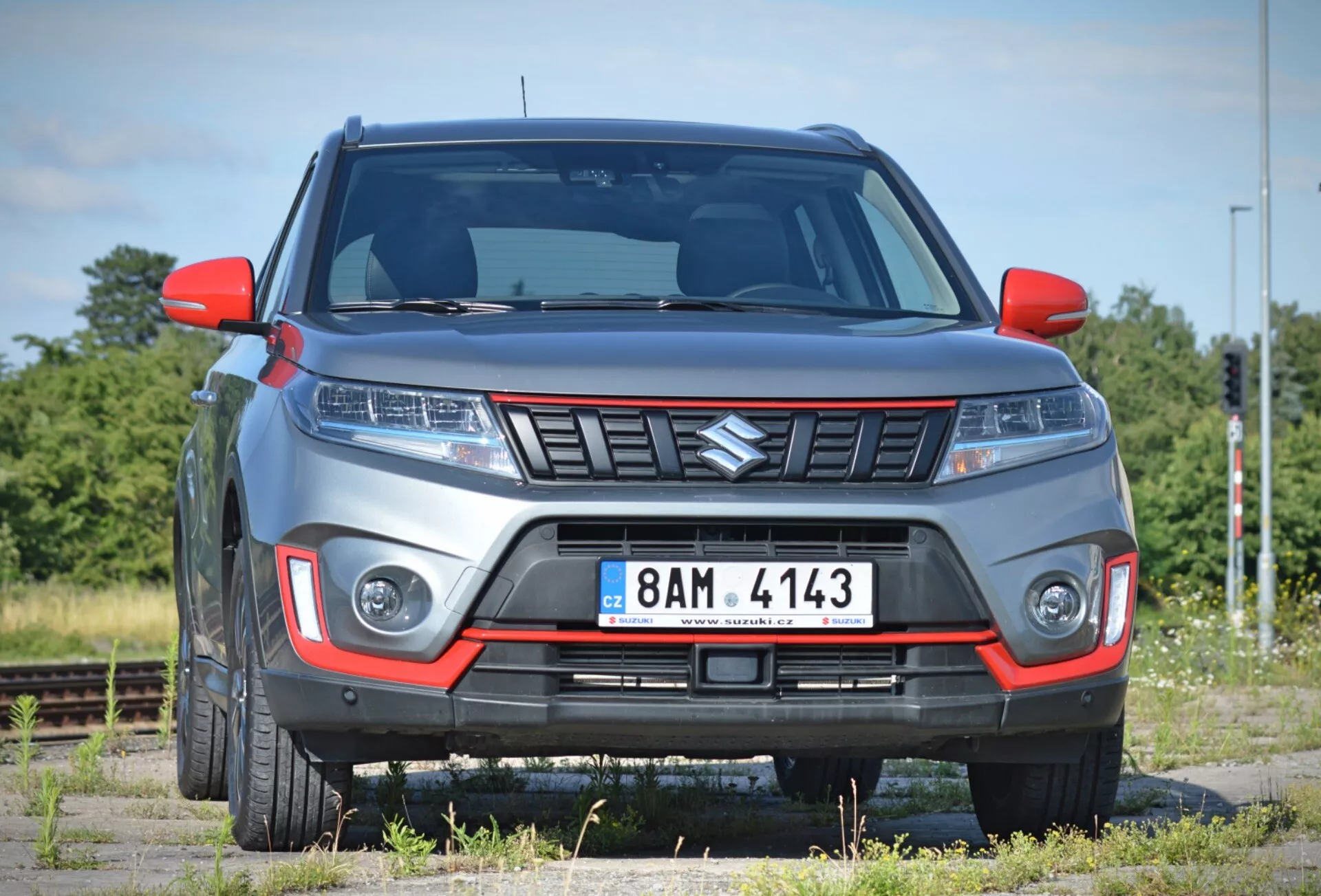 Obraz przedstawia nowoczesne SUV Suzuki Vitara 2018 pod lekkim kątem z przodu, ukazując przód i część pasażera na tle rzadkiego krajobrazu z torami kolejowymi. Samochód, zaparkowany na prawdopodobnie żwirowej nawierzchni, wyposażony jest w wyraźny grill o kanciastym kształcie, elegancki zestaw reflektorów prawdopodobnie z technologią LED lub HID oraz zintegrowane światła do jazdy dziennej lub przeciwmgielne w przednim zderzaku. Zderzak zawiera kontrastujący wstaw, sugerujący off-roadowy lub sportowy poziom wyposażenia, a widoczny prześwit podkreśla jego charakter SUV. Wzniesione linie na masce dodają agresywnego stylu, a boczne lusterka i nadkola wskazują na nowoczesny design. Częściowo widoczne alufelgi uzupełniają solidny wygląd. Zdjęcie wykonane z bliskiej odległości pozwala na szczegółowe zbadanie tych cech.