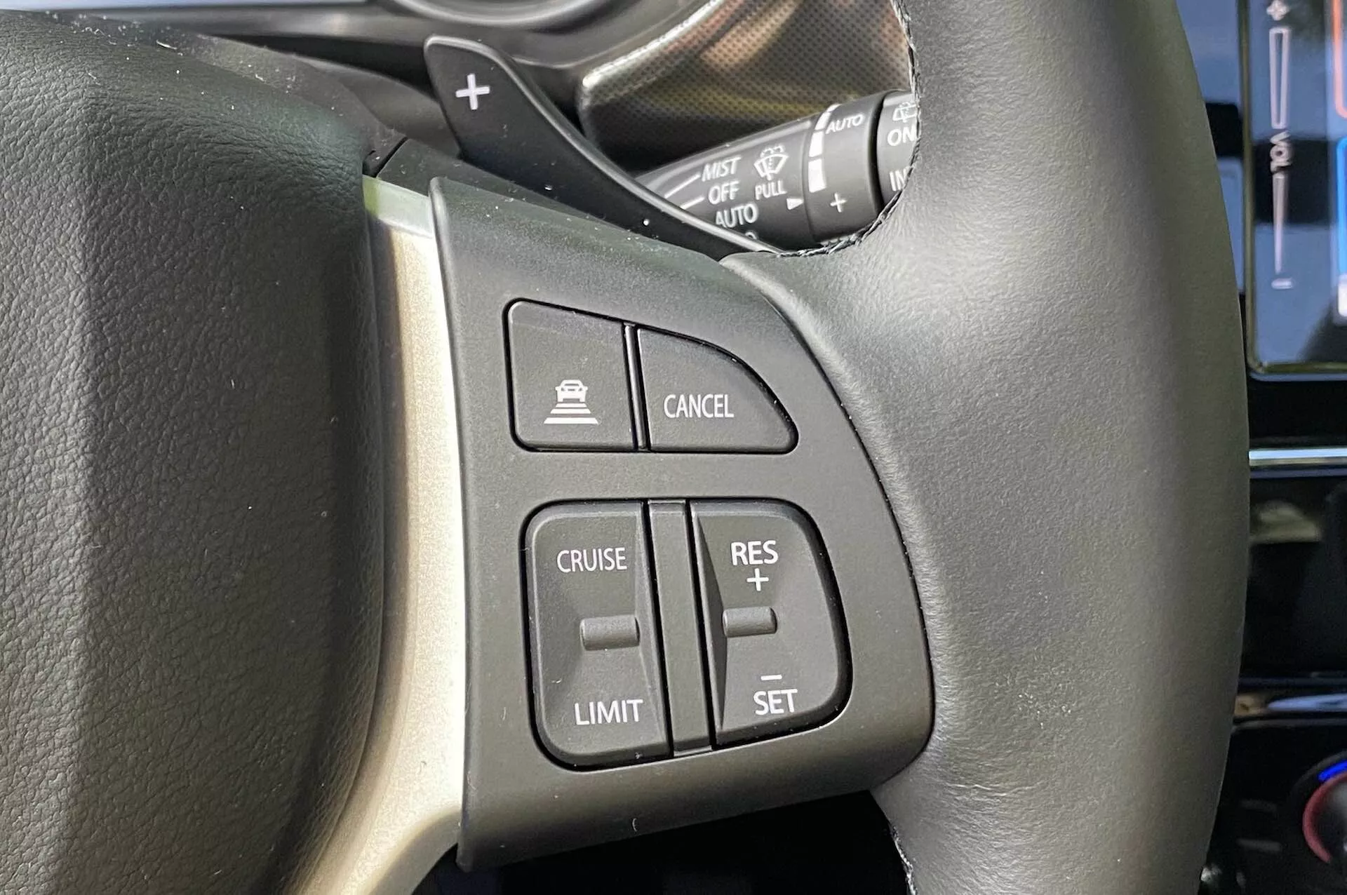 Zbliżenie na zamontowane na kierownicy przyciski w Suzuki Vitara 2018, ze szczególnym uwzględnieniem lewej strony. Czarna skórzana kierownica z widocznym przeszyciem posiada łatwo dostępne przyciski, w tym przycisk włączania tempomatu, przycisk włączania ogranicznika prędkości, przyciski ustawiania/doskonalenia tempomatu i przycisk anulowania. Na obrazku widać również dźwignię kontrolną prawdopodobnie do kierunkowskazów i świateł, widok z perspektywy kierowcy.