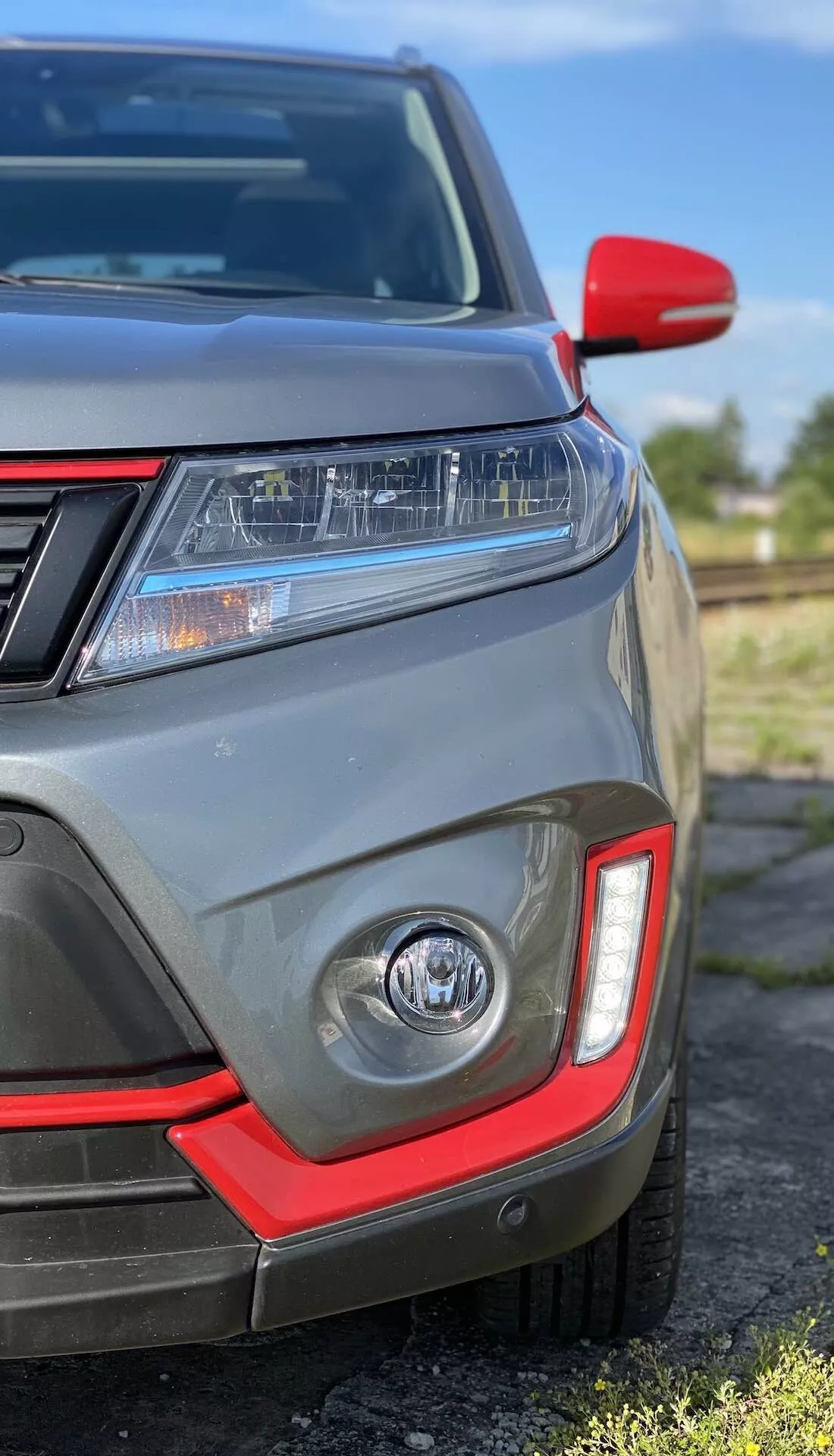 Zbliżenie na przedni lewy kwadrant Suzuki Vitara 2018 przedstawiające reflektory LED, światła do jazdy dziennej, reflektor przeciwmgielny z czerwonym akcentem, czujniki parkowania, częściowy grill przedni, lusterko boczne z czerwoną nasadką i metaliczny szary lakier.
