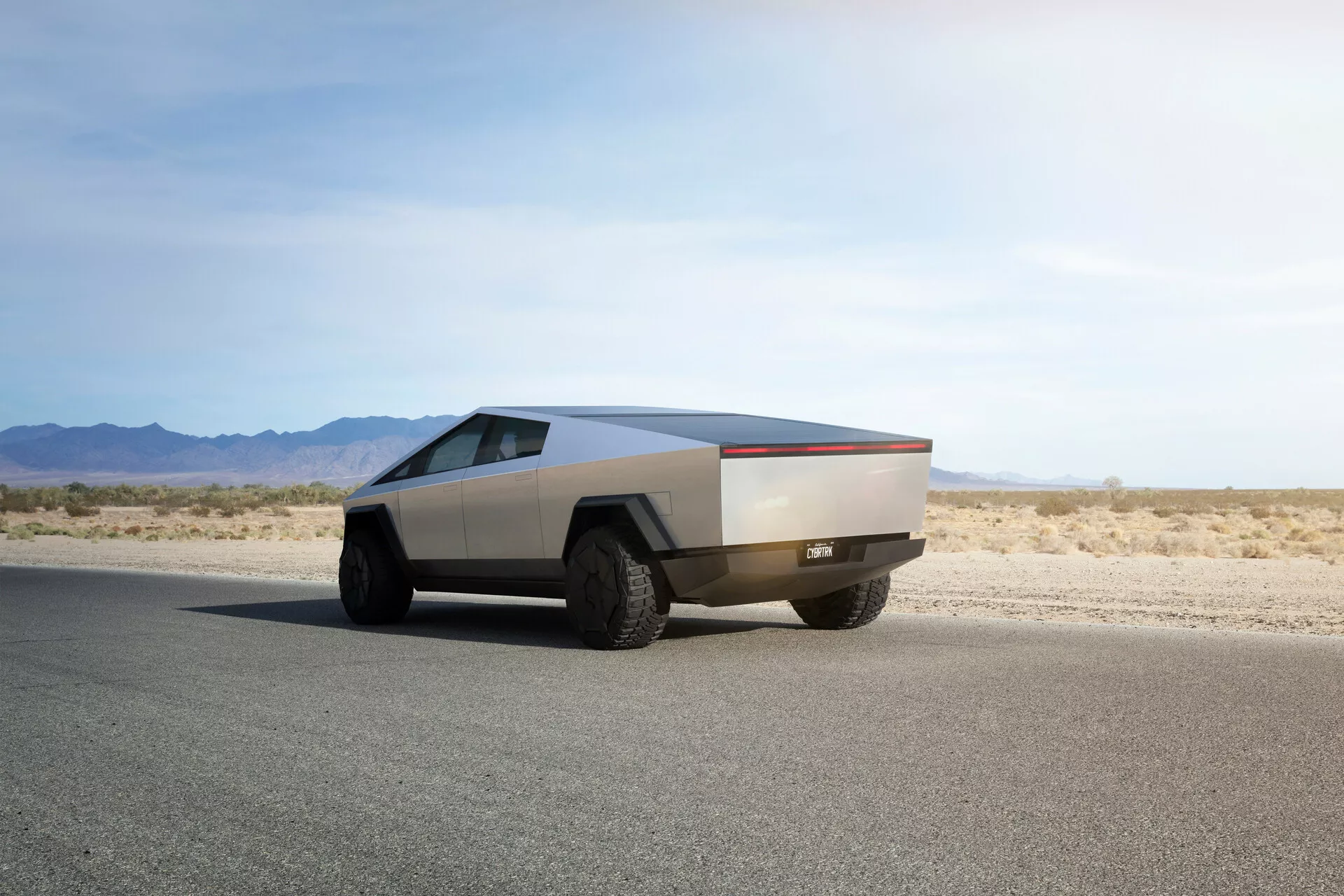 Futurystyczny Tesla Cybertruck 2021 na płaskiej asfaltowej drodze uchwycony pod kątem trzech czwartych od tylu, ukazujący charakterystyczny kanciasty design, metaliczne wykończenie, duże terenowe opony oraz brak tradycyjnych lusterek i klamek, co wskazuje na zaawansowane zautomatyzowane mechanizmy.