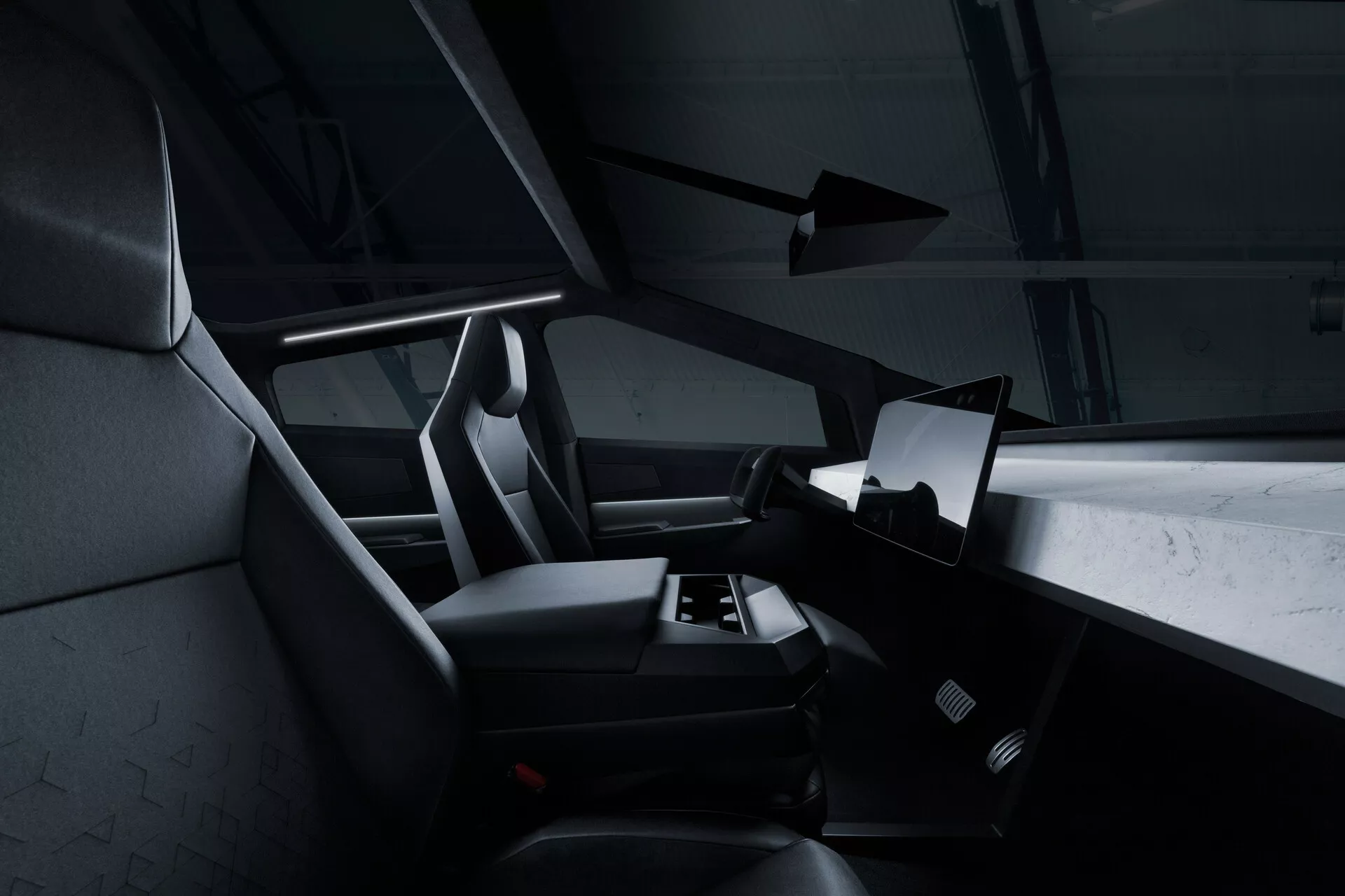 Widok wnętrza Tesla Cybertruck 2021 (1. generacja) z perspektywy tylnych siedzeń, pokazujący prawą stronę kabiny z dużym poziomym wyświetlaczem dotykowym dominującym w środkowej części deski rozdzielczej. Część kierownicy jest widoczna, ale głównie zasłonięta. Przednie siedzenia pasażera i kierowcy mają zintegrowane zagłówki i teksturowaną tapicerkę, prawdopodobnie z tkaniny lub skóry syntetycznej. W obszarze podłogi widać dywanik samochodowy, a brak widocznych ręcznych elementów sterujących sugeruje minimalistyczne, cyfrowe rozwiązanie. Ciemna paleta kolorów i subtelne oświetlenie akcentowe nadają nowoczesny, luksusowy wygląd.
