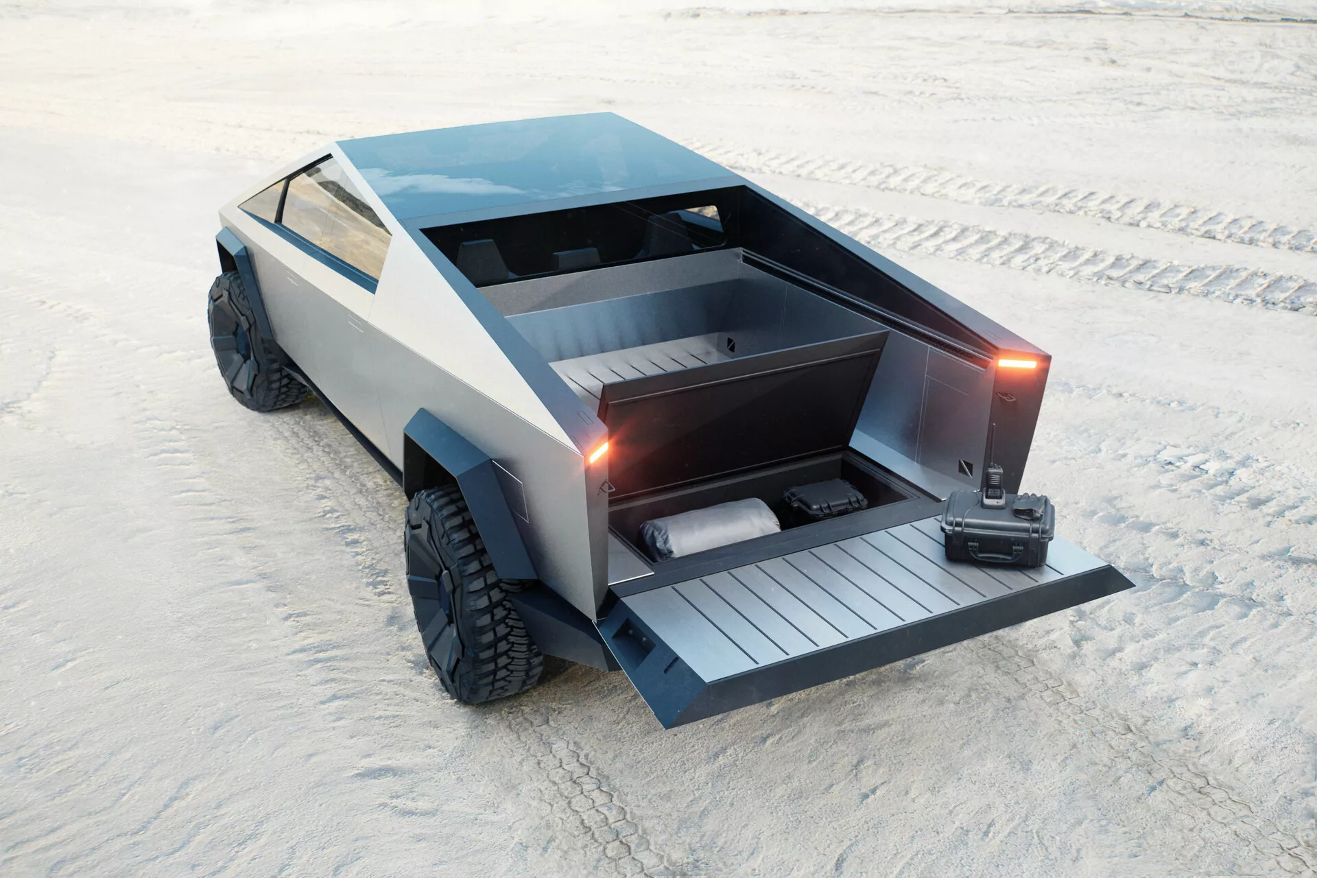 Futurystyczny Tesla Cybertruck 2021 (1. generacja) zaparkowany na piaszczystej powierzchni, uchwycony z podwyższonego tylno-trzyczwórkowego widoku. Kanciasty, wielościanowy design z plaskim bagażnikiem i złożoną, zdalnie sterowaną klapą, która odsłania wbudowaną rampę. Pojazd zawiera również blokowaną osłonę tonneau, duże terenowe opony i matowe szare wykończenie, co przyczynia się do jego minimalistycznej, przemysłowej estetyki. Brak tradycyjnych klamek drzwi ani bocznych lusterek, co sugeruje wysuwane klamki i lusterka oparte na kamerze. Podejrzewane jako elektryczne, z naciskiem na praktyczność i możliwości terenowe.