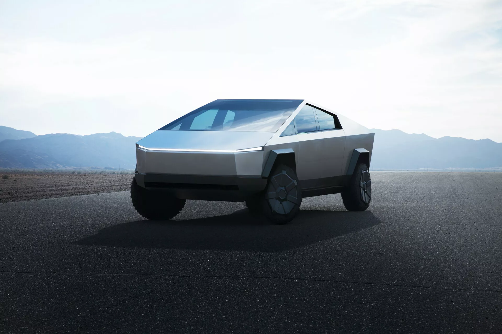 Zdjęcie Tesla Cybertruck 2021 (1. generacja) na drodze z równinami lub pustynnymi górami w tle w ciągu dnia i przy bezchmurnym niebie. Zdjęcie wykonane z niskiego kąta, pokazujące przód i bok ciężarówki, podkreślając jej kanciasty, futurystyczny design. Pojazd ma klinowaty przód, karoserię z płaskich paneli, cienkie poziome reflektory, ciągłą pochyłą szybę przednią, brak widocznych bocznych lusterek, duże koła terenowe z złożonymi felgami oraz brak przedniego grilla, co wskazuje na napęd elektryczny.