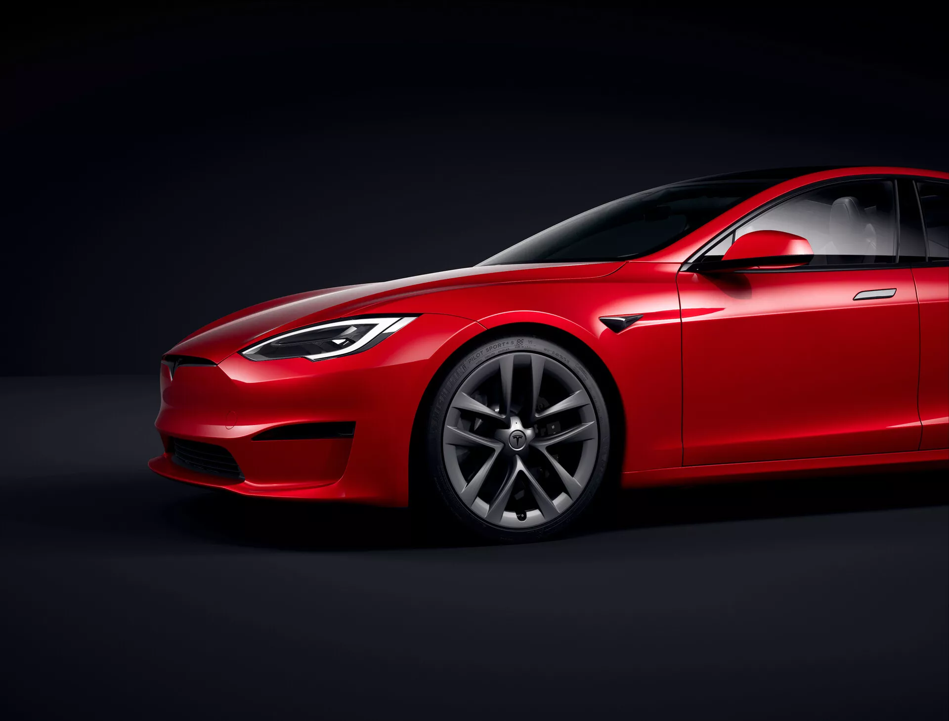 Tesla Model S 2016 (lift pierwszej generacji) czerwony sedan w studiu z kontrolowanym oświetleniem i czarnym tłem, niski kąt widzenia podkreślający aerodynamiczny profil i prześwit, widok trzy-czwarte z przodu pokazujący stronę kierowcy, reflektory, grill, część maski, wzór felg ze stopu premium z wieloszprychowym wzorem, przednią szybę z przyciemnianym szkłem, pochyloną linię dachu, lusterko boczne, klamki drzwi i linię okien harmonijnie wkomponowaną w elegancki design, podkreślającą wykończenie lakieru, linie konturowe i rzeźbione panele sugerujące luksus i osiągi, skoncentrowane oświetlenie pokazujące szczegóły designu.
