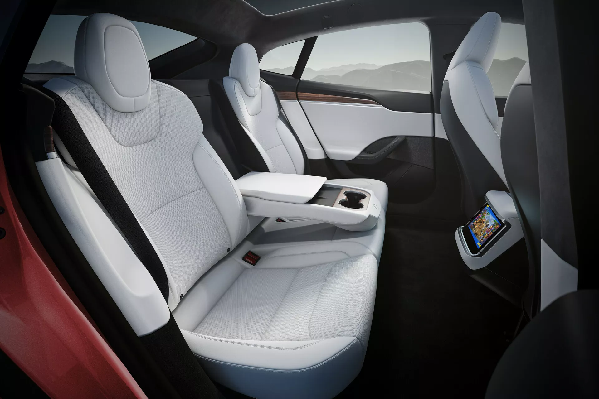 Wnętrze Tesla Model S 2016 (1. generacji facelift) prezentujące luksusowe, białe skórzane fotele z czarnymi akcentami, ergonomiczny design i dużo miejsca na nogi. Zdjęcie uchwyca siedzenia po prawej stronie z nieco odgórnej, pochylonej perspektywy, podkreślając funkcjonalną konsolę centralną kończącą się przed tylnymi siedzeniami, wyposażoną w ekran dotykowy dla pasażerów tylnych i bezprzewodowe ładowanie smartfonów. Panoramiczny szklany dach oferuje naturalne światło i poczucie przestronności.