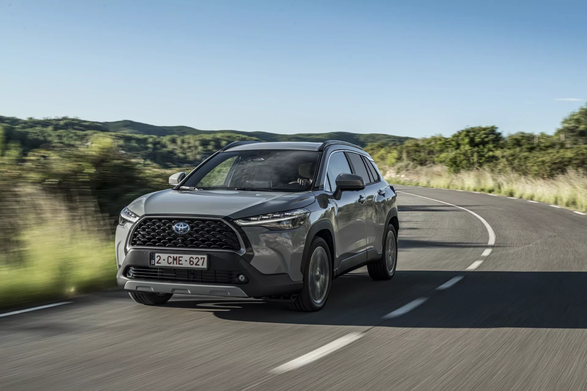 Model Toyota Corolla Cross 1. generacji z 2022 roku w ruchu na wiejskiej drodze, prezentujący widok z przodu i boku, uwypuklający LEDowe reflektory, przedni grill z emblematem i boczne felgi aluminiowe