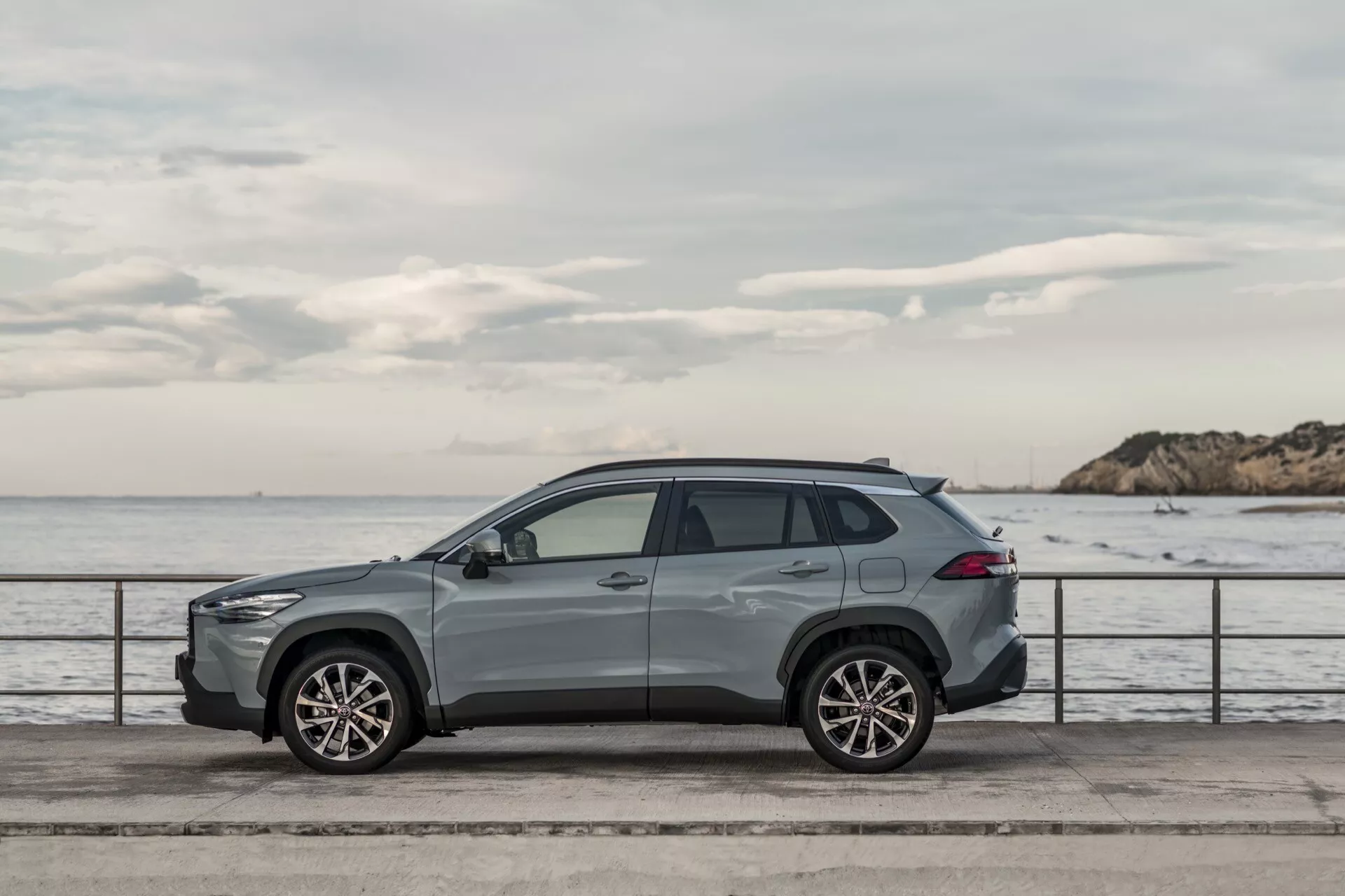 Toyota Corolla Cross 1. generacji 2022 zaparkowana równolegle do promenady przy wodzie, prezentująca swój aerodynamiczny profil, elegancki design, felgi aluminiowe o wielu ramionach, relingi dachowe i wykończenie z wyższej półki w spokojnym otoczeniu z wodą i pochmurnym niebem w tle