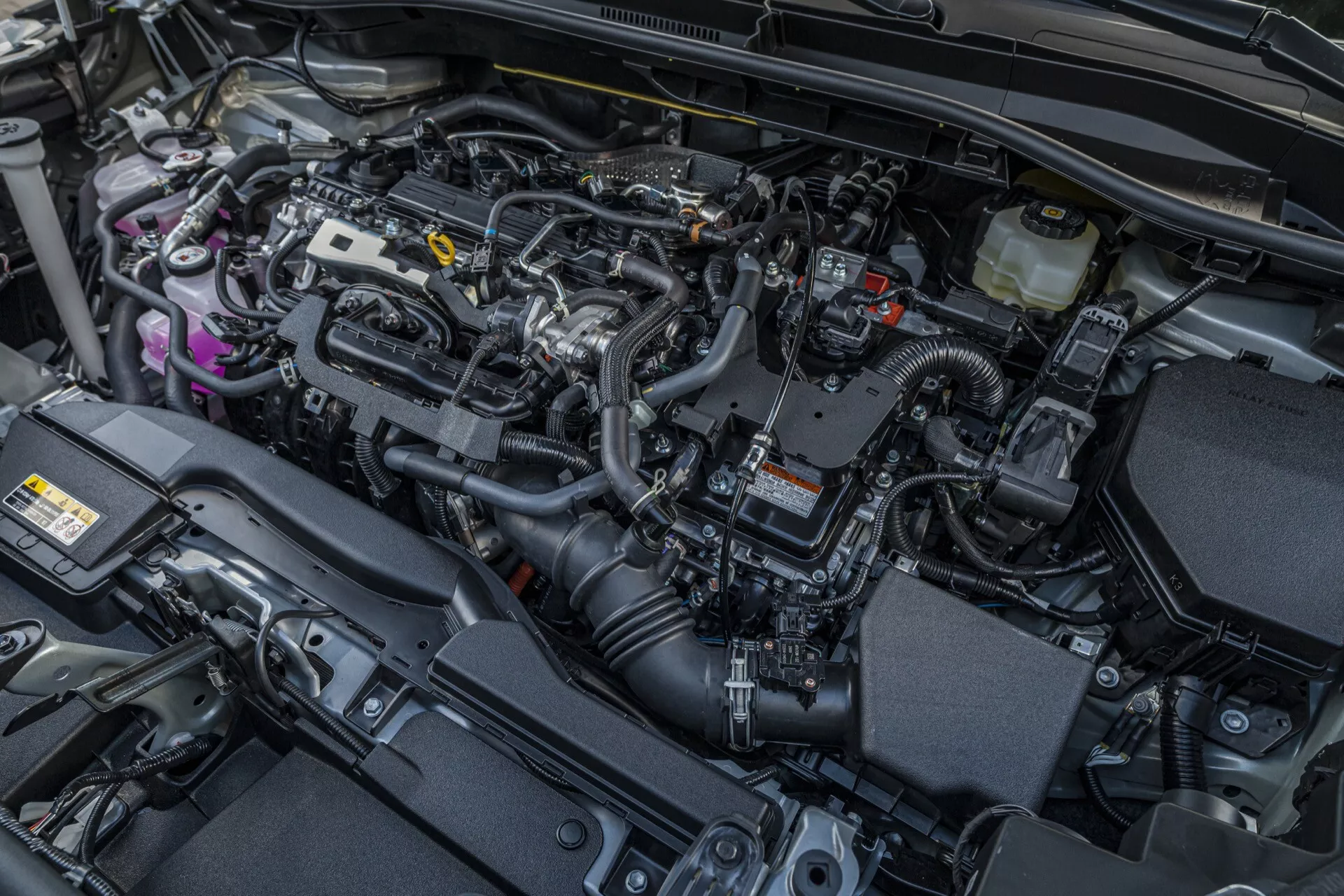 Szczegółowy widok komory silnika Toyota Corolla Cross 2022 (1. generacja) podkreślający jej nowoczesny silnik spalinowy i różnorodne technologie samochodowe, takie jak elektroniczny system zarządzania silnikiem, pokrywa silnika tłumiąca hałas, zbiorniki płynów, system dolotu powietrza i strategiczne komponenty zarządzania ciepłem.