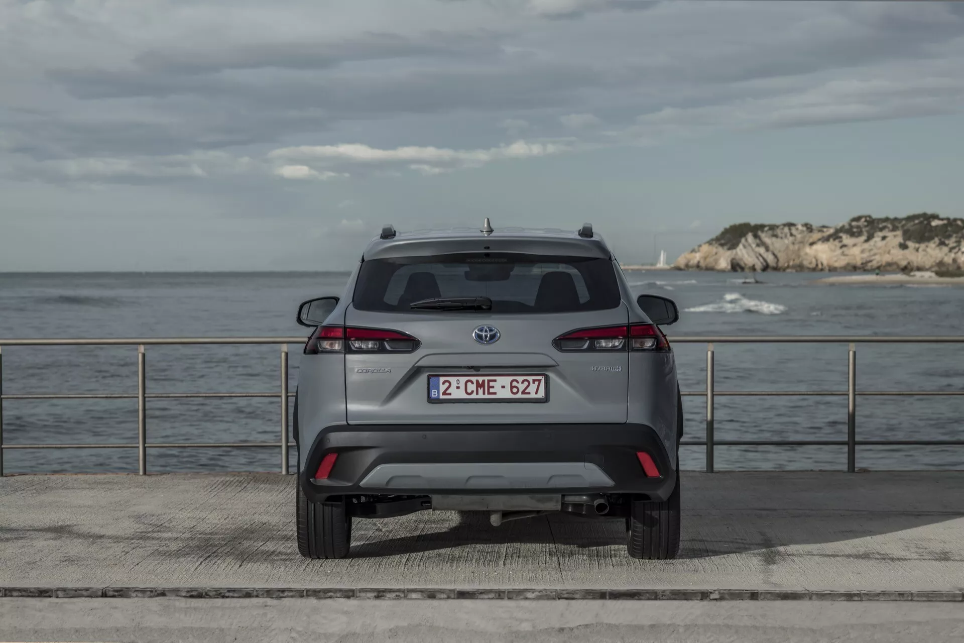 Tylny widok Toyoty Corolla Cross 2022 zaparkowanego na platformie nad morzem z naturalnym oświetleniem, widokiem na horyzont, pełnymi światłami, tylną szybą, wycieraczką, emblematem marki, tylnym zderzakiem, odznaką modelu, tablicą rejestracyjną oraz technologicznymi elementami bezpieczeństwa jak czujniki parkowania.