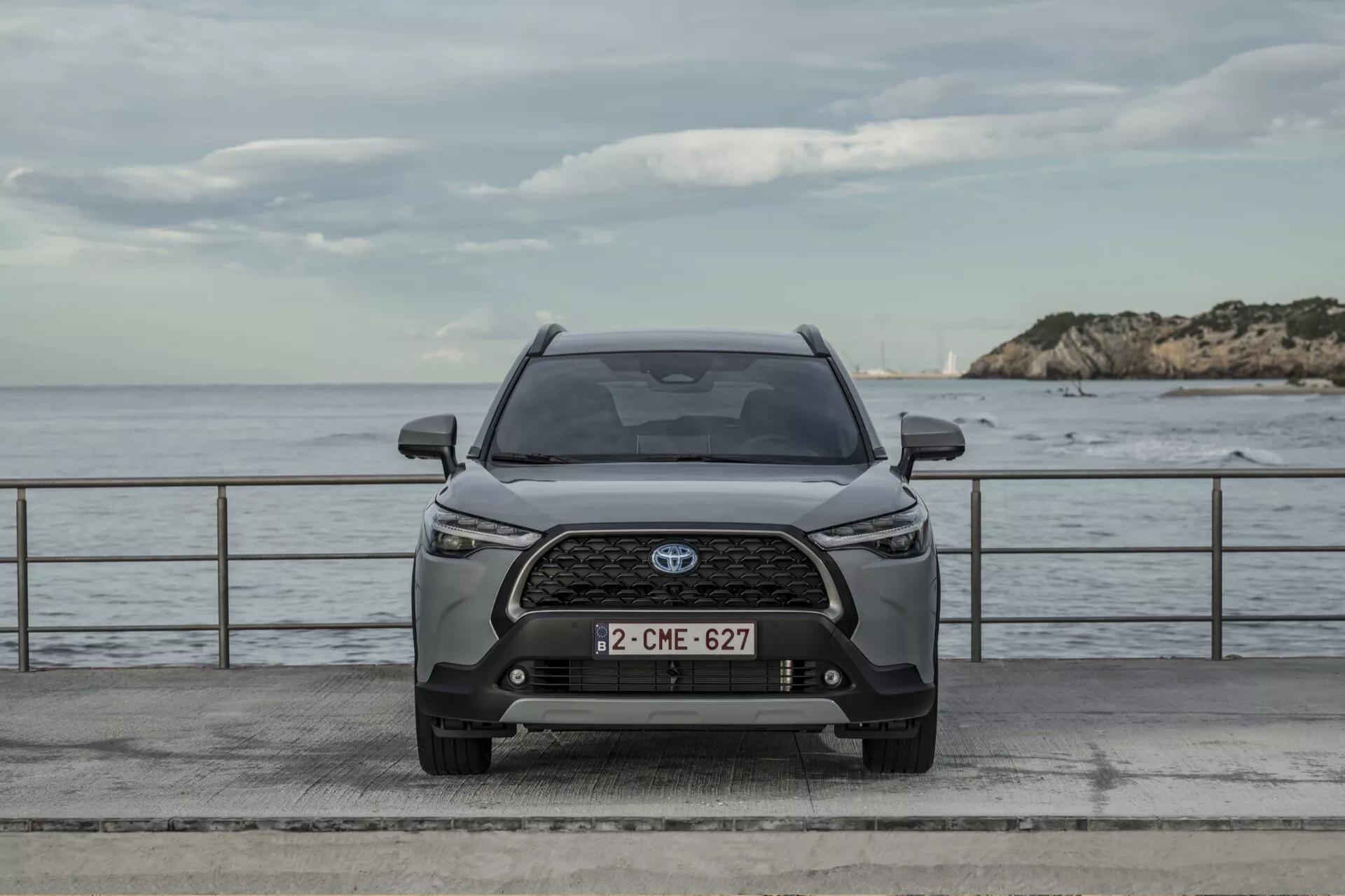 Przedni widok Toyota Corolla Cross 2022 pokazujący symetryczny design, zaawansowane reflektory LED, złożoną kratkę z emblematem Toyota i nowoczesne cechy zewnętrzne na neutralnym tle z wodą i pochmurnym niebem