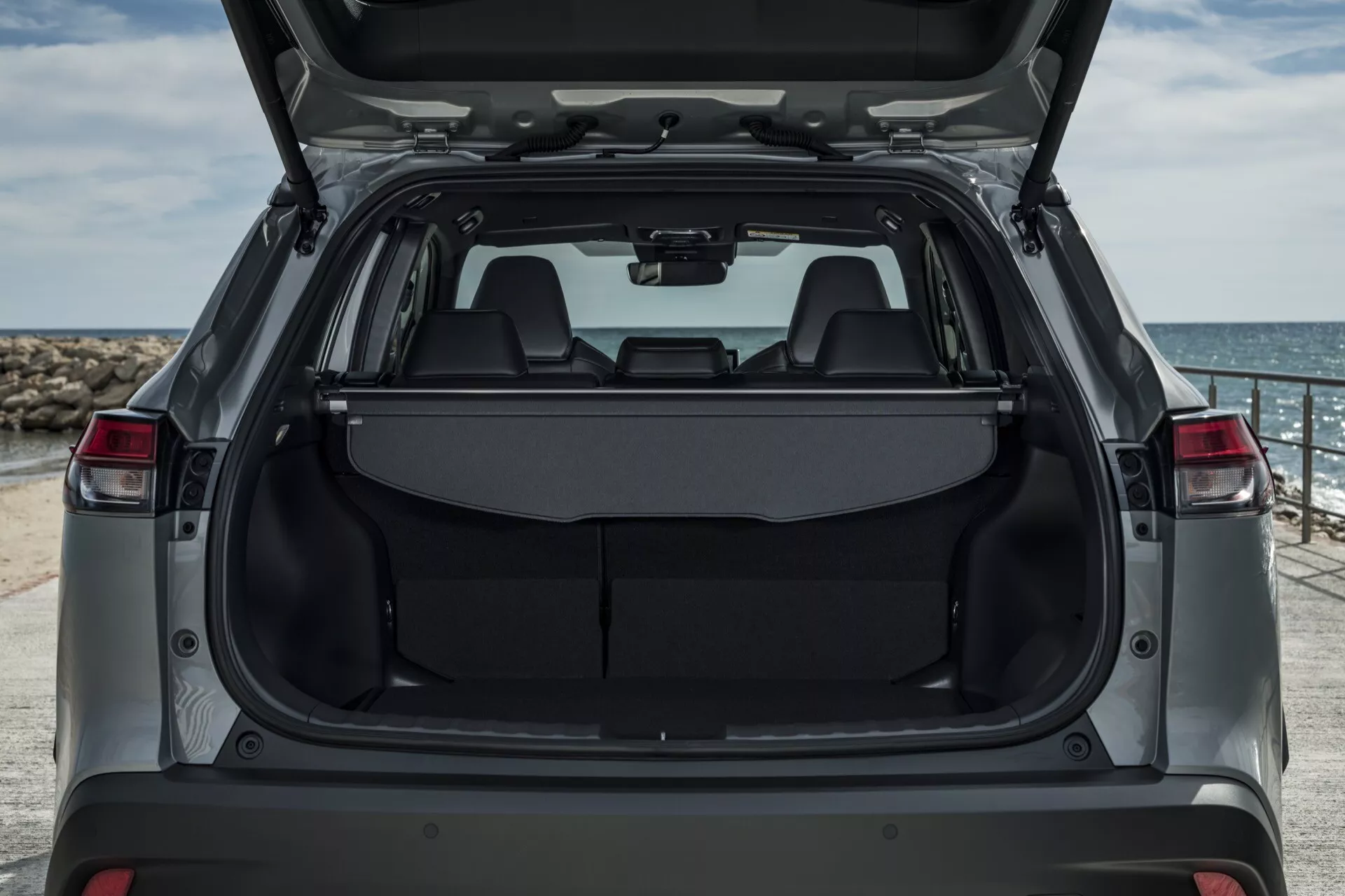 Toyota Corolla Cross pierwszej generacji z 2022 roku prezentująca swój tylny wnętrze przez otwartą klapę bagażnika, z rozkładanymi tylnymi siedzeniami, wykładziną bagażową z hakami i chowaną osłoną bagażu, na tle krajobrazu nadmorskiego