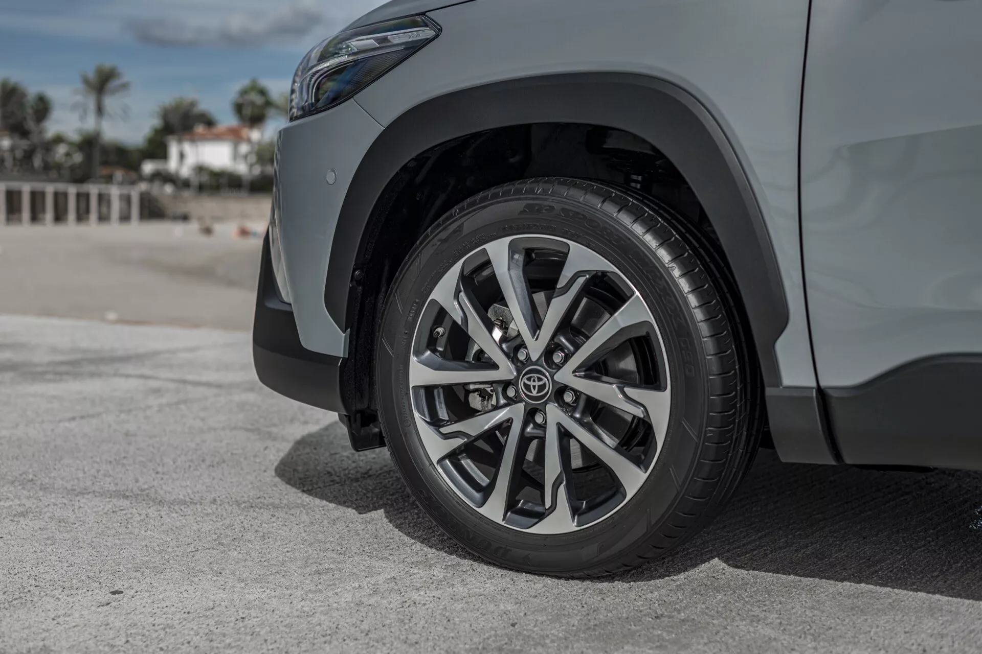 Zbliżenie na przednie prawe koło i błotnik Toyota Corolla Cross 2022, z wielopałkowym designem obręczy i znaczkiem marki, na tle palm i niebieskiego nieba