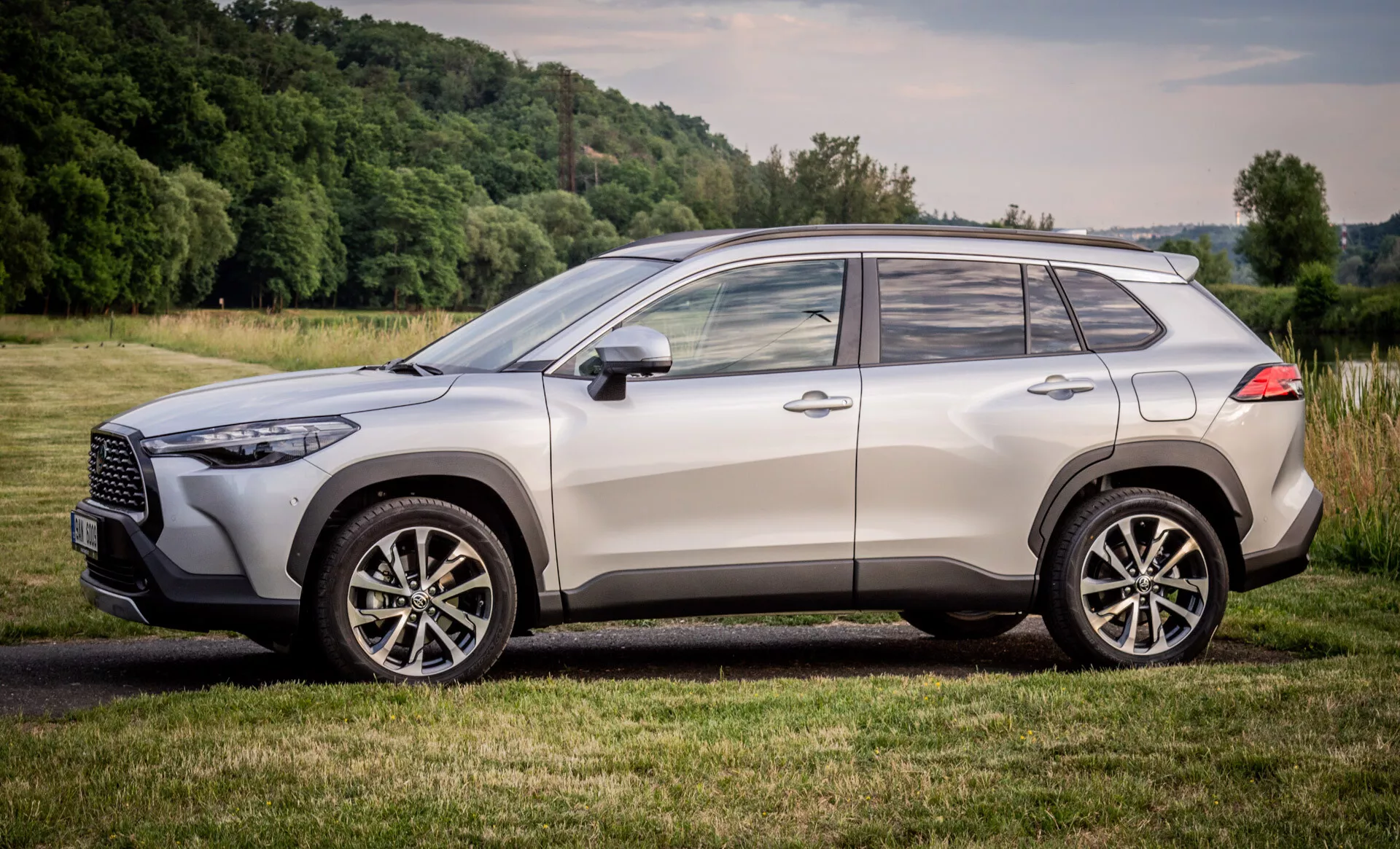 Toyota Corolla Cross 2022 zaparkowana na zewnątrz na trawiastym terenie, przedstawia profil boczny, nowoczesny design SUV z akcentami chromowanymi, felgami aluminiowymi i zaawansowaną technologią oświetlenia
