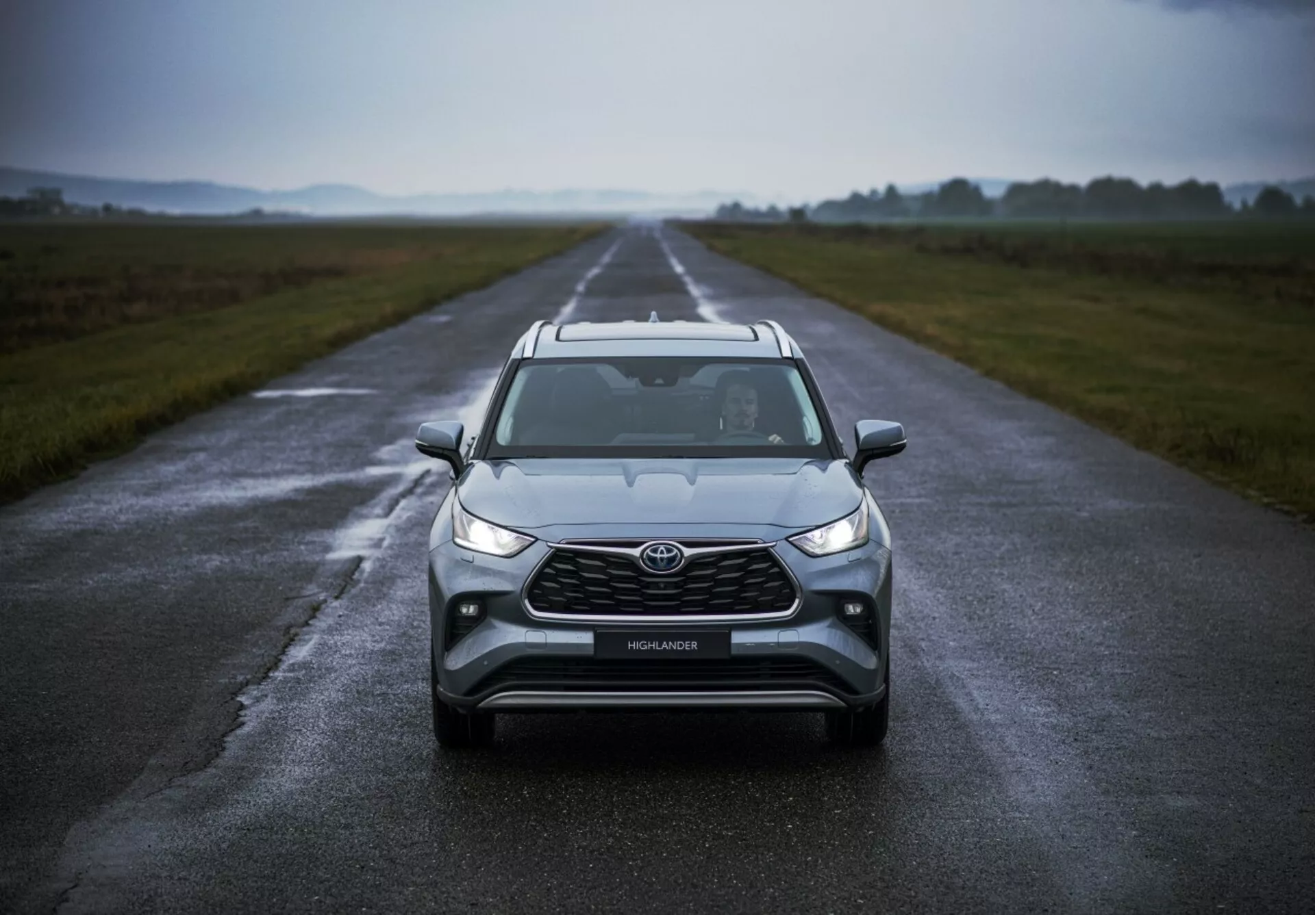 Toyota Highlander 2021 umiejscowiony na mokrej drodze podkreślający jego frontowy design, pochmurna pogoda, z reflektorami LED, światłami przeciwmgielnymi, dużą atrapą i zaawansowaną technologią bezpieczeństwa