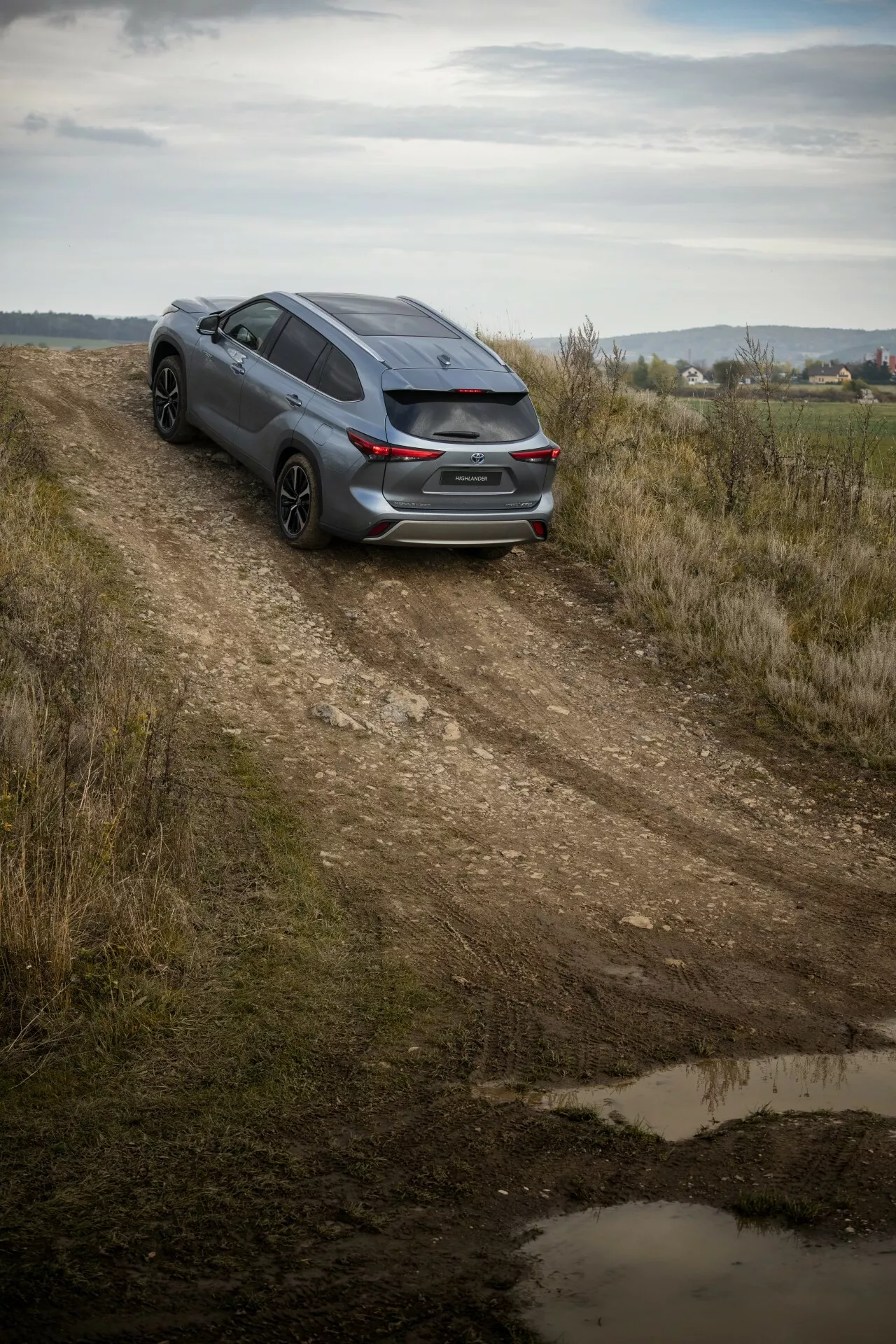 Toyota Highlander 2021 w ciemnym kolorze uchwycona z wysokiego kąta na drodze gruntowej, prezentuje widok z tyłu trzech kwartałów, sportowy design i potencjał do lekkiej jazdy w terenie wśród wiejskiego krajobrazu