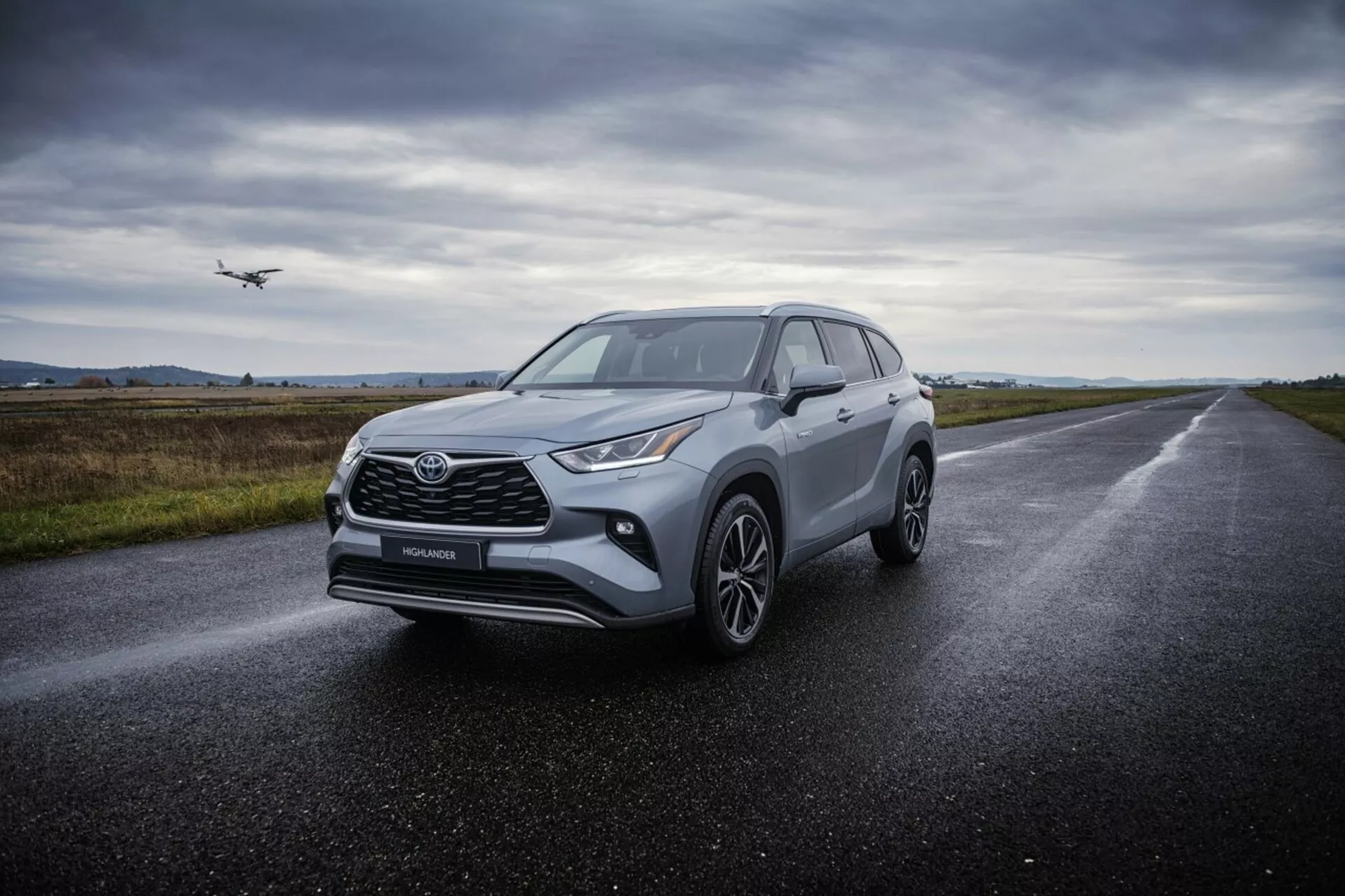 Crossover SUV Toyota Highlander 2021 na mokrej asfaltowej drodze, prezentujący kątowe reflektory, wyrazistą przednią kratę, wieloramienne koła ze stopu i aerodynamiczny design pod rozproszonym zachmurzonym oświetleniem