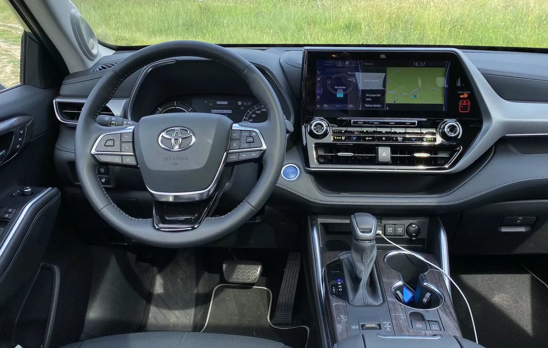 Wnętrze modelu Toyota Highlander 2.5 Hybrid Business Edition 2021 z perspektywy kierowcy. Obraz pokazuje kierownicę z emblematem Toyoty, wielofunkcyjne przyciski, częściowo widoczny zestaw wskaźników oraz konsolę środkową z ekranem dotykowym systemu infotainment wyświetlającym mapę nawigacyjną. Widoczne są również sterowanie klimatem, przegródka na połowę, elektroniczna dźwignia zmiany biegów i hamulec postojowy, podkreślając nowoczesny design i technologiczne rozwiązania pojazdu.