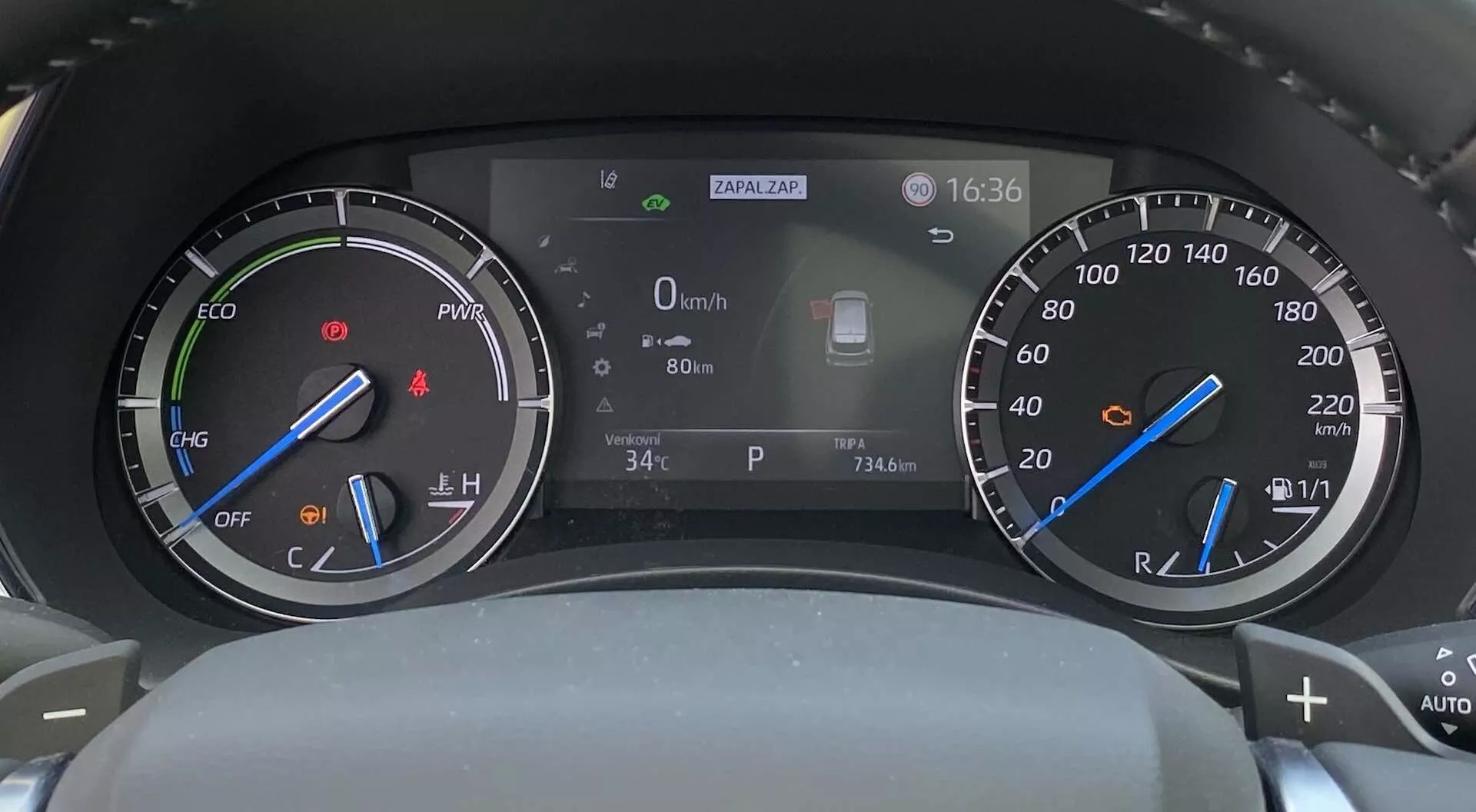 Hybrydowy deska rozdzielcza cyfrowo-analogowa Toyota Highlander 2021, widziana z siedzenia kierowcy, z obrotomierzem z wskaźnikiem trybu ECO po lewej stronie, prędkościomierzem z wskaźnikiem poziomu paliwa po prawej stronie i centralnym wyświetlaczem cyfrowym pokazującym prędkość (0 km/h), temperaturę zewnętrzną (34°C), zegar (16:36) oraz ikonę systemu wspomagania kierowcy.