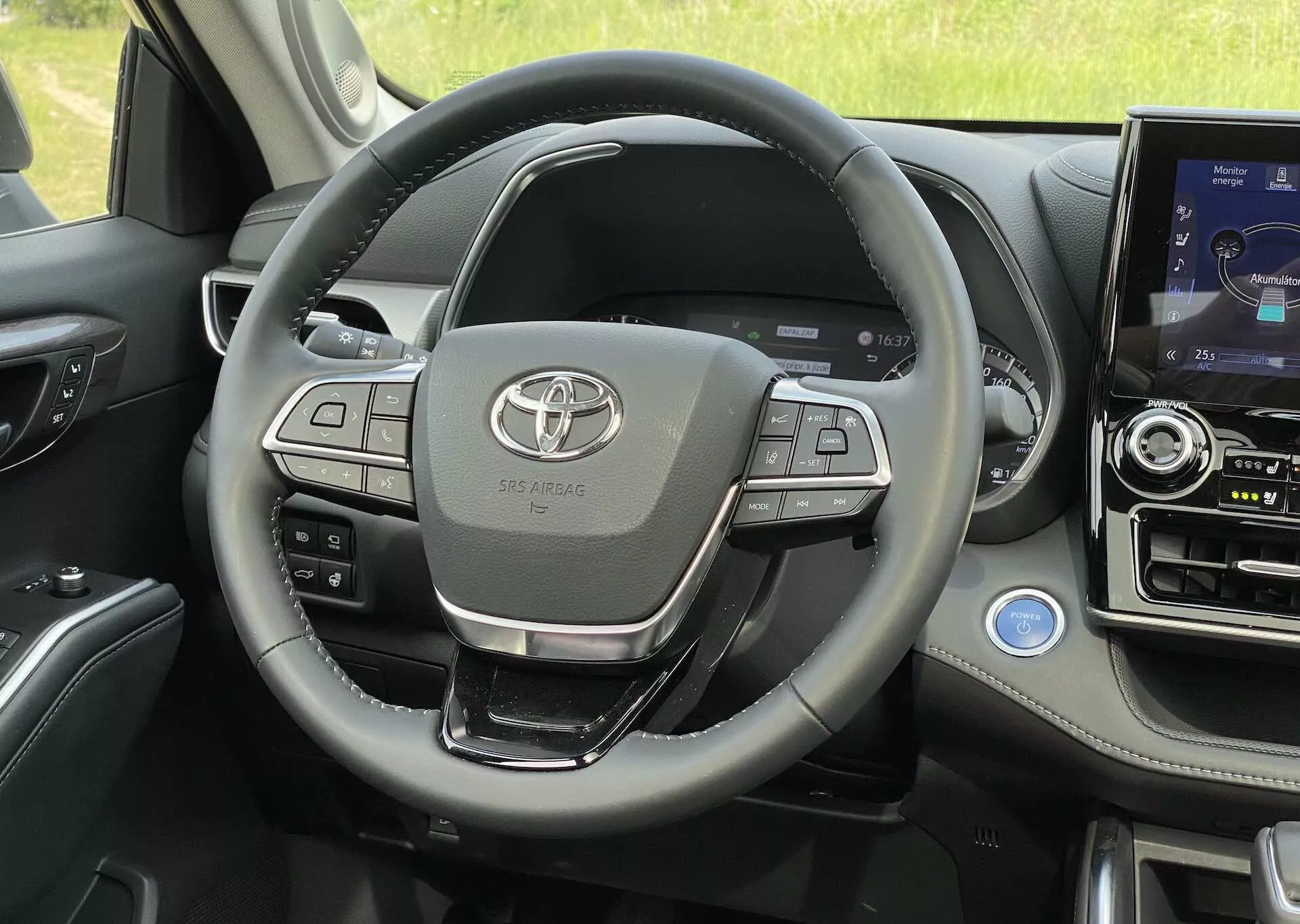 Wnętrze Toyota Highlander 2.5 Hybrid Business Edition 2021 widziane z perspektywy pierwszoosobowej, z naciskiem na wielofunkcyjną kierownicę z przyciskami do sterowania dźwiękiem, łączności telefonicznej i tempomatu, z wyraźnym emblematem Toyota na środku. Widoczna deska rozdzielcza, centralna konsola z dotykowym systemem infotainment, dźwignia zmiany biegów, panel sterowania drzwiami i wentylacja, ukazujące materiały wysokiej jakości i zaawansowane funkcje.