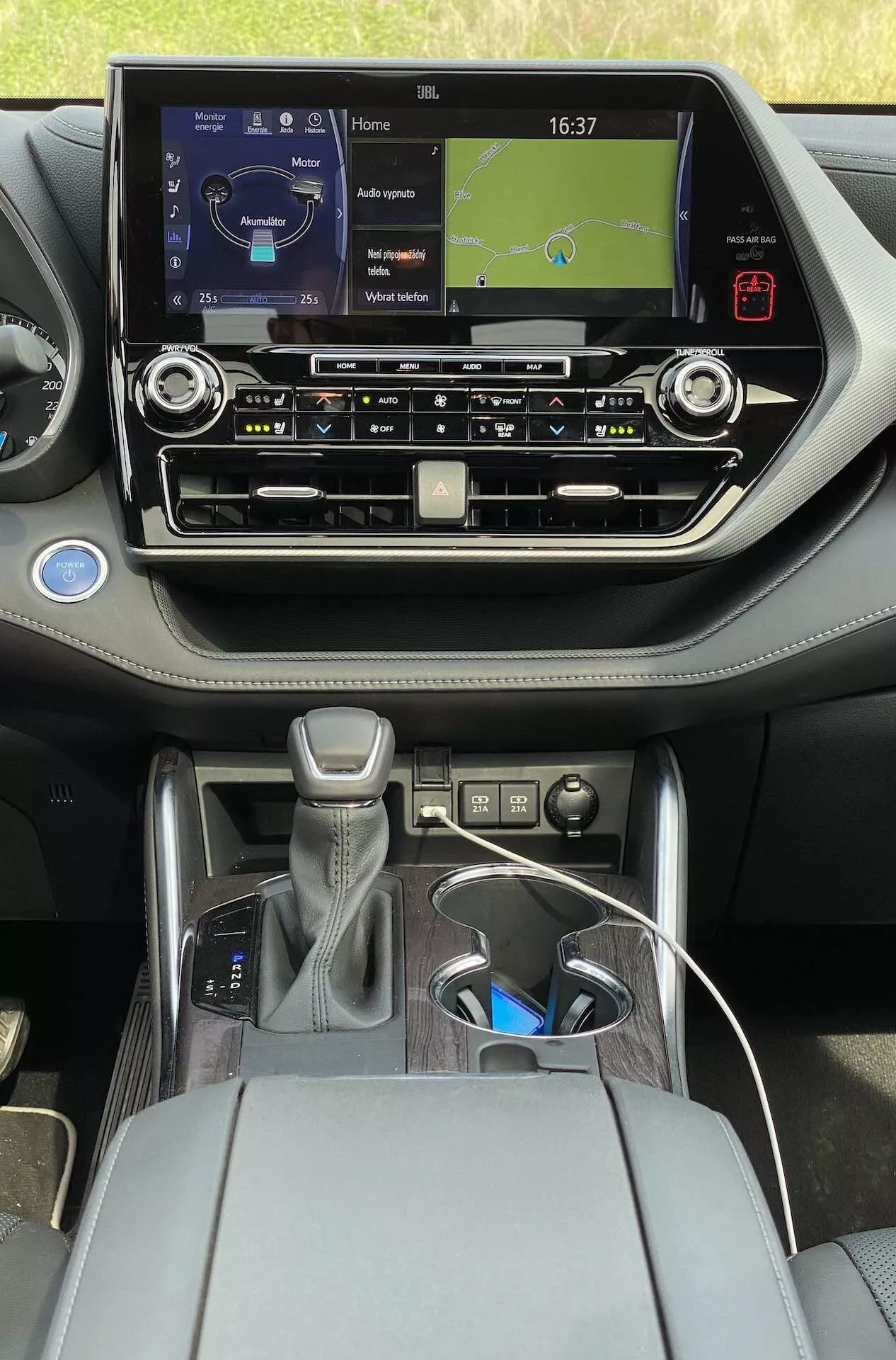 Widok wnętrza Toyota Highlander 2.5 Hybrid Business Edition 2021 z tylnego siedzenia, z nowoczesnym dotykowym systemem infotainment z zegarkiem i mapą nawigacyjną, przyciskami fizycznymi do kontroli klimatu, wentylacją, automatyczną skrzynią biegów z osłoną ze skóry i srebrnymi akcentami, pokrętłem sterującym, centralnym podłokietnikiem z otwartą przestrzenią do przechowywania, dwoma uchwytami na napoje, kablem USB do ładowania i współczesnym, dwukolorowym wykończeniem z metalicznymi akcentami.