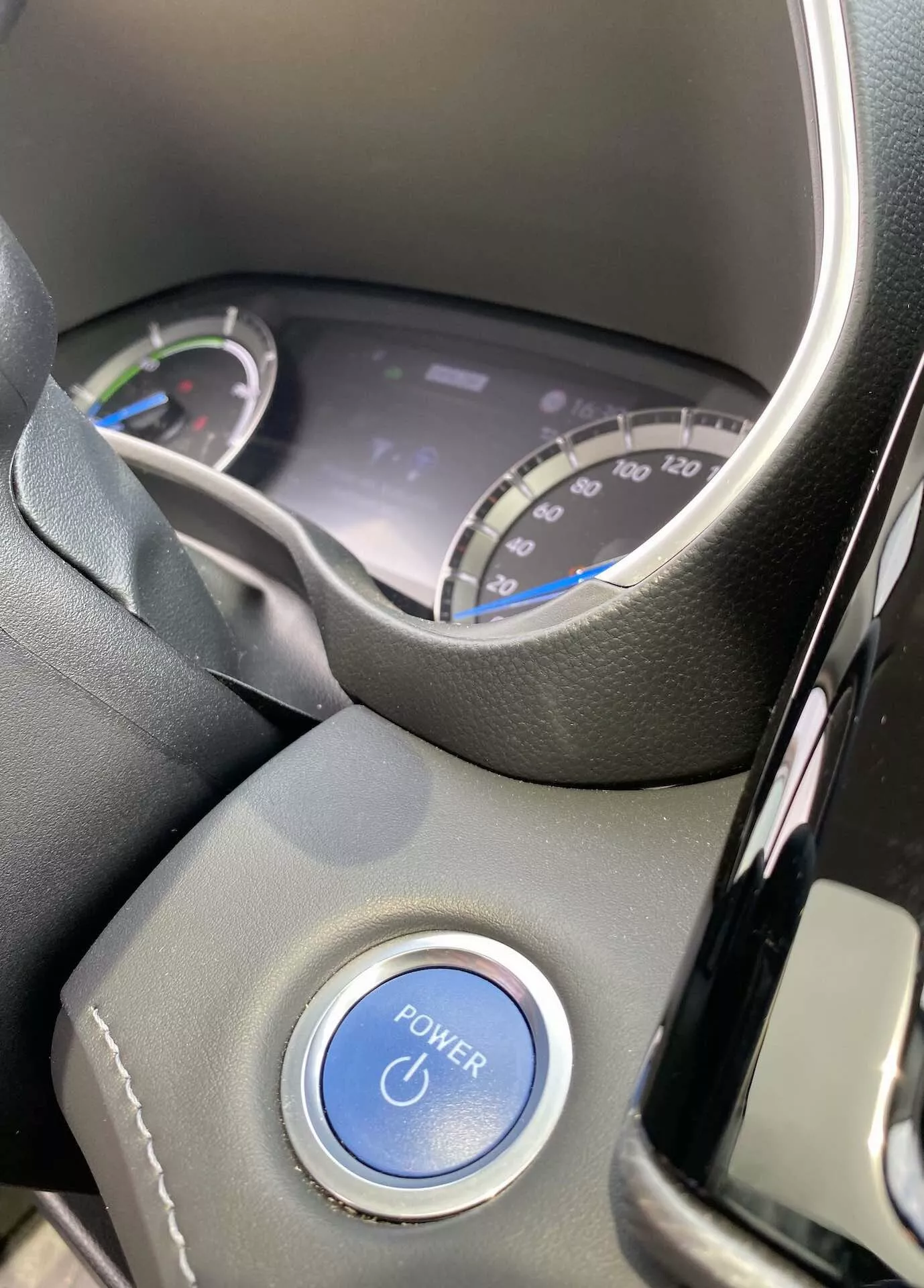 Widok wnętrza Toyoty Highlander 2021 2.5 Hybrid Business Edition z perspektywy kierowcy. Obraz pokazuje część kierownicy i cyfrowy zestaw wskaźników przez górną część kierownicy. Zestaw zawiera prędkościomierz oznaczony do 220 km/h oraz wskaźnik zużycia energii, co sugeruje hybrydową naturę pojazdu. Na kolumnie kierownicy widoczny jest charakterystyczny niebieski przycisk 'POWER', podkreślający system zapłonu przyciskiem. Wnętrze ma nowoczesne czarne tworzywa sztuczne i kierownicę z skóry lub podobnego materiału, z naturalnymi warunkami świetlnymi podkreślającymi nowy lub dobrze utrzymany stan samochodu.