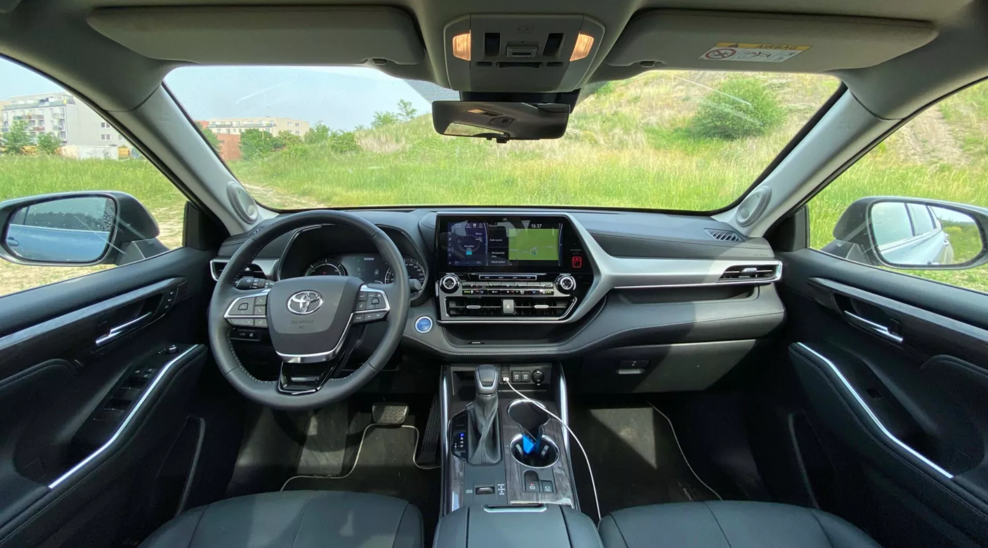 Widok wnętrza Toyota Highlander 2021 Hybrid Business Edition z tylnego siedzenia, prezentujący symetryczną perspektywę przedniego kokpitu. Kierownica, z emblematem pojazdu i zamontowanymi przyciskami audio, znajduje się wyraźnie po lewej stronie. Centralny panel deski rozdzielczej posiada dotykowy ekran infotainment bez przycisków fizycznych, uzupełniony przyciskami i pokrętłami do sterowania klimatyzacją poniżej. Dźwignia zmiany biegów, nowoczesna dźwignia elektroniczna, jest widoczna w dolnej części zdjęcia, w pobliżu potencjalnej strefy ładowania smartfonów. Luksusowe wnętrze charakteryzuje się połączeniem skórzanych foteli, miękkimi materiałami na desce rozdzielczej i chromowanymi akcentami. Szyberdach wpuszcza naturalne światło, a panele sterujące znajdują się w pobliżu lusterka wstecznego. Na zewnątrz widać krajobraz naturalny przez przednie okna.