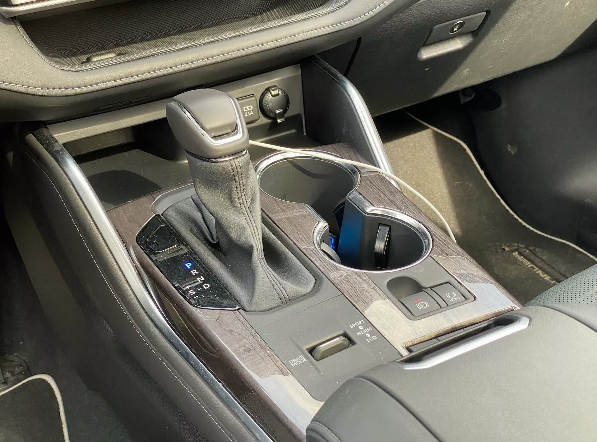 Widok wnętrza Toyoty Highlander 2.5 Hybrid Business Edition 2021, koncentrujący się na konsoli środkowej, na której znajduje się dźwignia zmiany biegów automatycznych ze skórzanymi przeszyciami, kontrolki trybu jazdy, uchwyty na kubki, przyciski sterujące, schowek w konsoli centralnej z wykończeniem drewnianym oraz częściowo widoczne regulatory fotela.