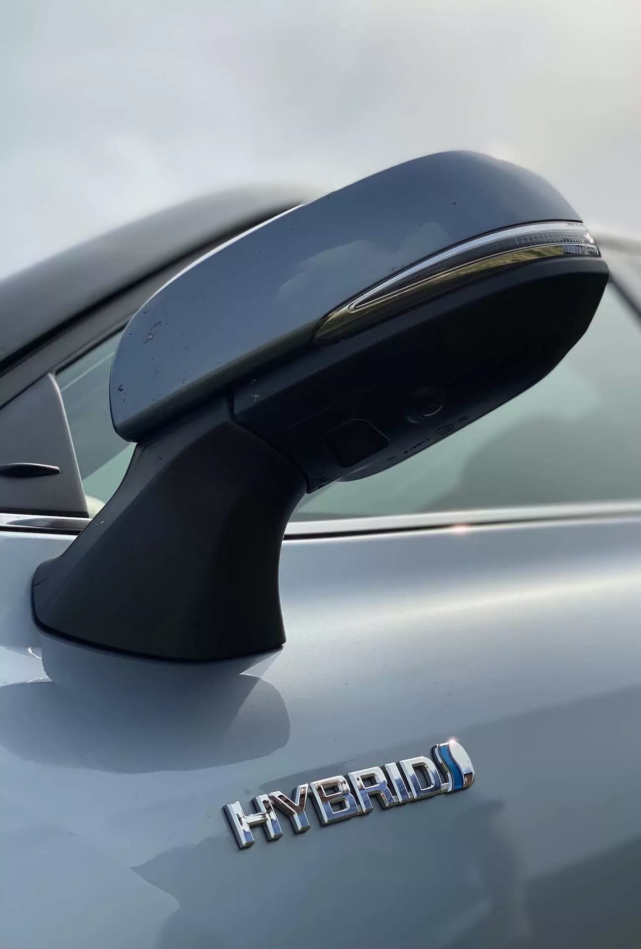 Zbliżenie na lusterko zewnętrzne od strony kierowcy i częściowy widok boczny Toyoty Highlander 2021 Hybrid Business Edition. Lusterko, zamontowane na drzwiach, ma aerodynamiczny kształt i zintegrowane światło kierunkowskazu. Zdjęcie wykonane z niskiego kąta podkreśla napis 'HYBRID' oznaczający ekologiczny napęd hybrydowy. Metaliczny szary lakier samochodu jest wyraźnie widoczny. Szczegóły są wyraźne dzięki małej głębi ostrości, która rozmywa tło.