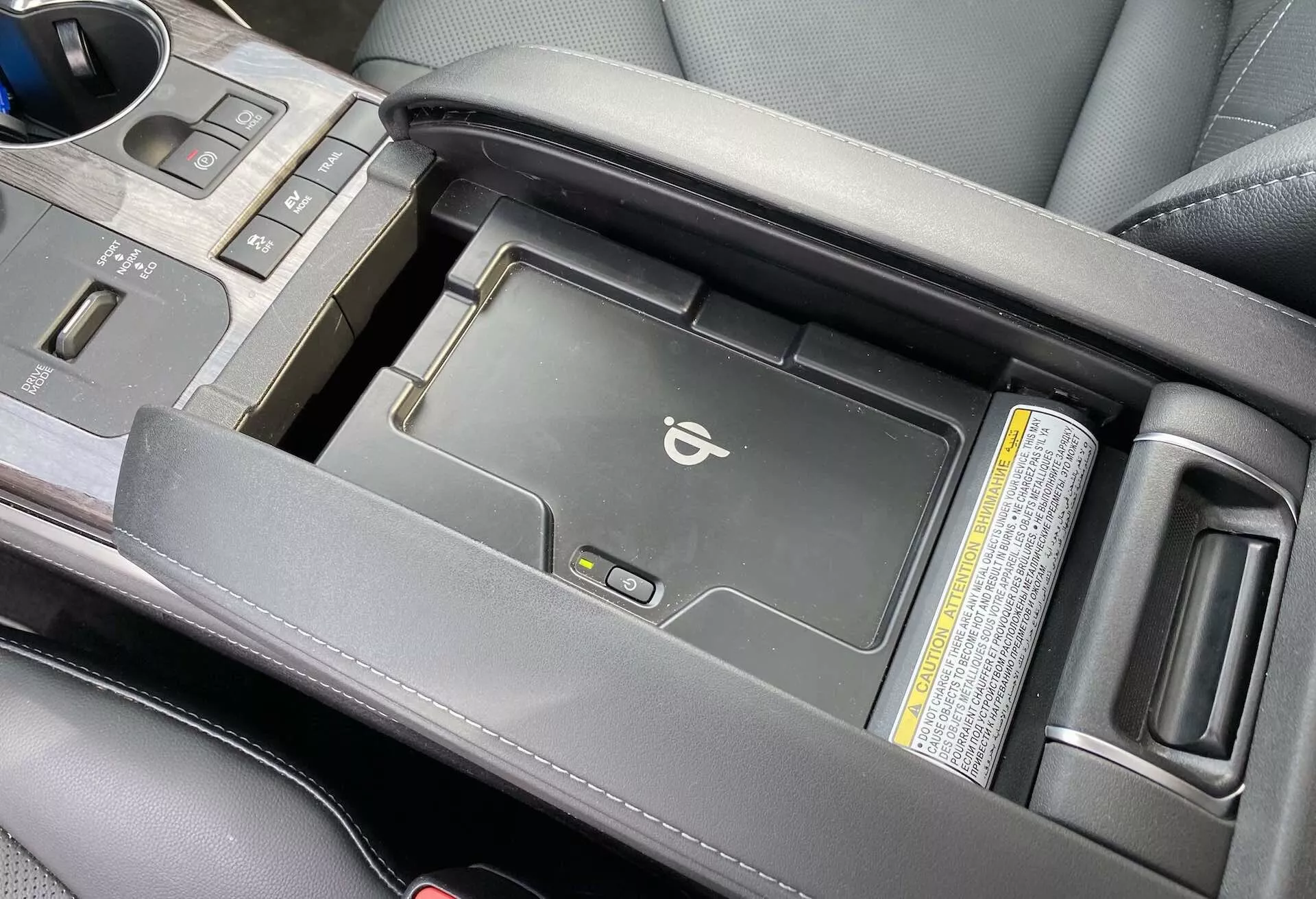 Zbliżenie konsoli centralnej modelu Toyota Highlander 2021 Hybrid Business Edition z nieco podwyższonego kąta, ukazujące częściowo otwarty obszar centralnego podłokietnika, z wbudowaną bezprzewodową ładowarką na urządzenia mobilne. Obraz uwydatnia luksusowy czarny skórzany podłokietnik, dźwignię zmiany biegów i uchwyty na kubki, skupiając się na nowoczesnych wygodach technologicznych oraz jakości estetycznej.