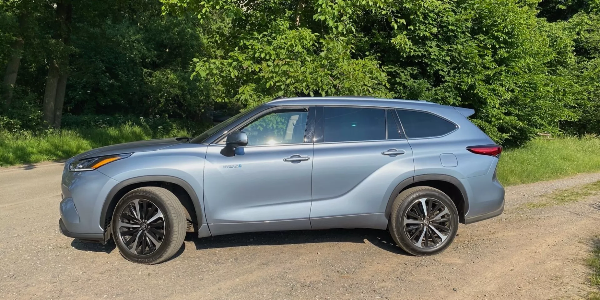 Boczny profil crossovera SUV Toyota Highlander 2.5 Hybrid Business Edition z 2021 roku z karoserią w metalicznym srebrze, zaparkowanego na powierzchni żwirowej lub gruntowej, otoczonego zieloną roślinnością w parku naturalnym lub na wiejskim terenie. Pojazd charakteryzuje się nowoczesnym, aerodynamicznym designem z liniami konturowymi i eleganckim, opływowym kształtem okien. Lusterka boczne są dopasowane do koloru nadwozia, a skomplikowane, wieloszprychowe felgi wyposażone są w niskoprofilowe opony. Relingi dachowe dodają praktyczności i solidnego stylu, widoczne klamki drzwi łączą się z linią charakterystyczną biegnącą przez nie, a samochód emanuje poczuciem ruchu naprzód, nawet gdy stoi.