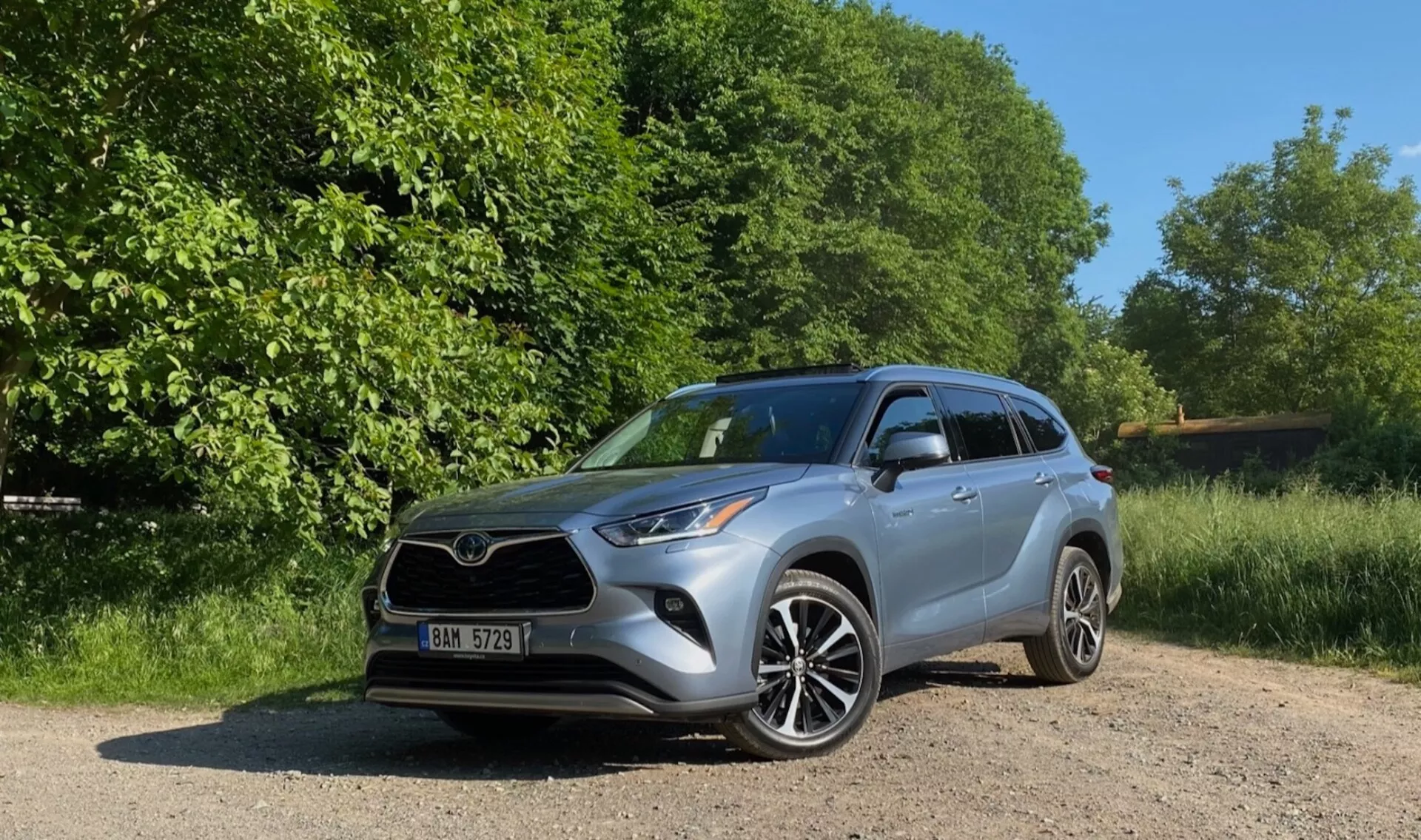 Srebrny Toyota Highlander 2.5 Hybrid Business Edition z 2021 roku jest zaparkowany na żwirowej ścieżce otoczonej bujnymi drzewami i krzewami. Dobrze oświetlony słoneczny dzień podkreśla szczegółowe elementy designu pojazdu, w tym reflektory LED, aluminiowe koła z podwójnymi ramionami i zintegrowane kierunkowskazy w lusterkach. Zdjęcie jest wykonane z niskiego kąta w widoku z przedniego trzy czwarte, co podkreśla solidną postawę SUV, przedni grill, reflektory i profil boczny.