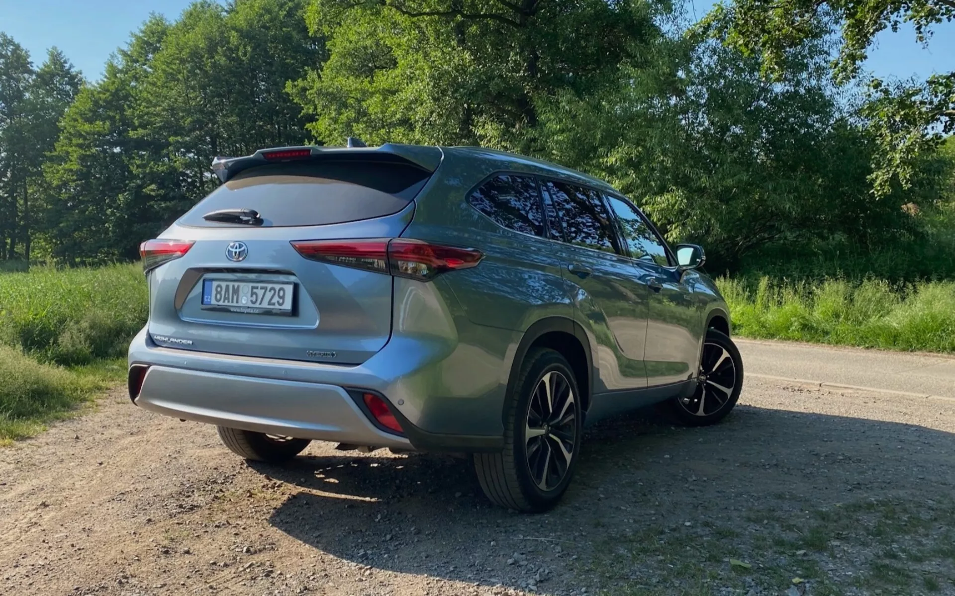 Widok tylnego trzy czwarte SUV-a Toyota Highlander 2021 2.5 Hybrid Business Edition, uchwycony od poziomu ziemi pod kątem w górę. Zdjęcie podkreśla prawą tylną stronę, w tym tylny zderzak, tylne światło, klapę z tylnym spojlerem dachowym, drzwi pasażera, okno i tylne koło. Metaliczny lakier SUV-a lśni w słońcu, ustawiony na nierównej, nieutwardzonej powierzchni otoczonej zielenią.