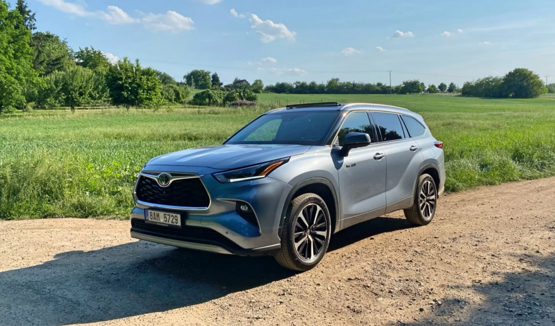 Obraz SUV Toyota Highlander 2.5 Hybrid Business Edition 2021 zrobiony z widoku z trzech czwartych frontu na drodze szutrowej z roślinnością i czystym niebem w tle. Zdjęcie przedstawia przedni grill, reflektory, maskę, przednie błotniki, drzwi kierowcy, lusterka boczne, koła i relingi dachowe pojazdu. Ma nowoczesne aluminiowe felgi, reflektory LED lub projektorowe, lusterka boczne w kolorze nadwozia z potencjalnymi zintegrowanymi kierunkowskazami, szyberdach i przyciemniane szyby. SUV jest zaprojektowany z wyższym prześwitem i praktycznymi elementami jak światła przeciwmgłowe i wloty powietrza w przednim zderzaku.
