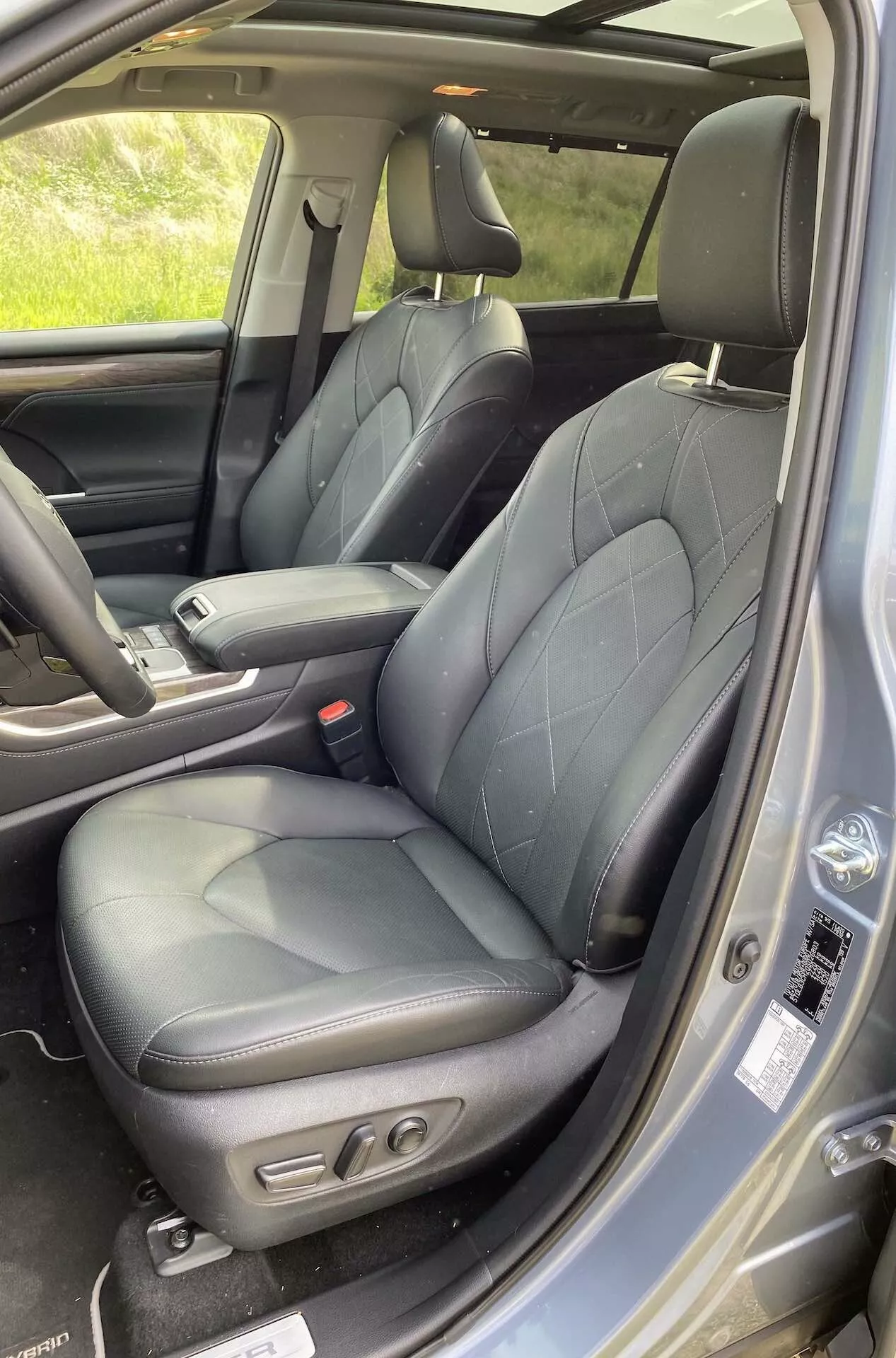 Wnętrze Toyota Highlander 2021 2.5 Hybrid Business Edition, ukazujące obszar siedzenia kierowcy z pikowaną czarną skórzaną tapicerką, bocznymi wzmocnieniami i elektrycznymi elementami sterującymi. Widoczna jest część konsoli centralnej, panel drzwi z przyciskami do obsługi elektrycznych szyb i lusterek, kratką głośnika oraz pas bezpieczeństwa przedniego siedzenia. Luksusowy i ergonomiczny design jest podkreślony naturalnym światłem.