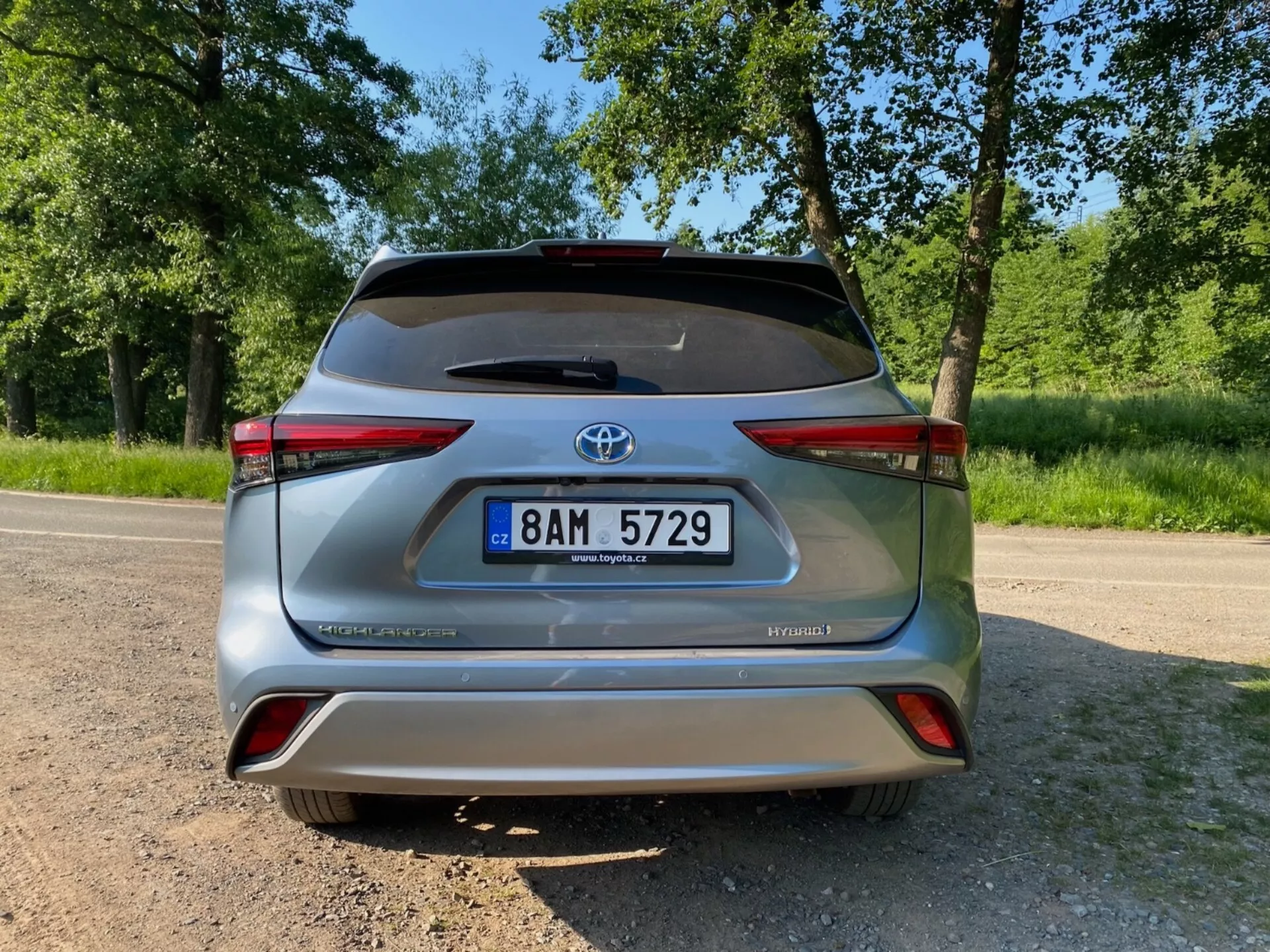 Widok tylu srebrnego Toyota Highlander 2.5 Hybrid Business Edition z widocznymi oznaczeniami HYBRID i AWD-i, centralnym numerem rejestracyjnym, emblematem Toyota oraz tylnymi światłami z elementem odblaskowym, wszystko na asfaltowej nawierzchni z zielonymi liśćmi w tle.