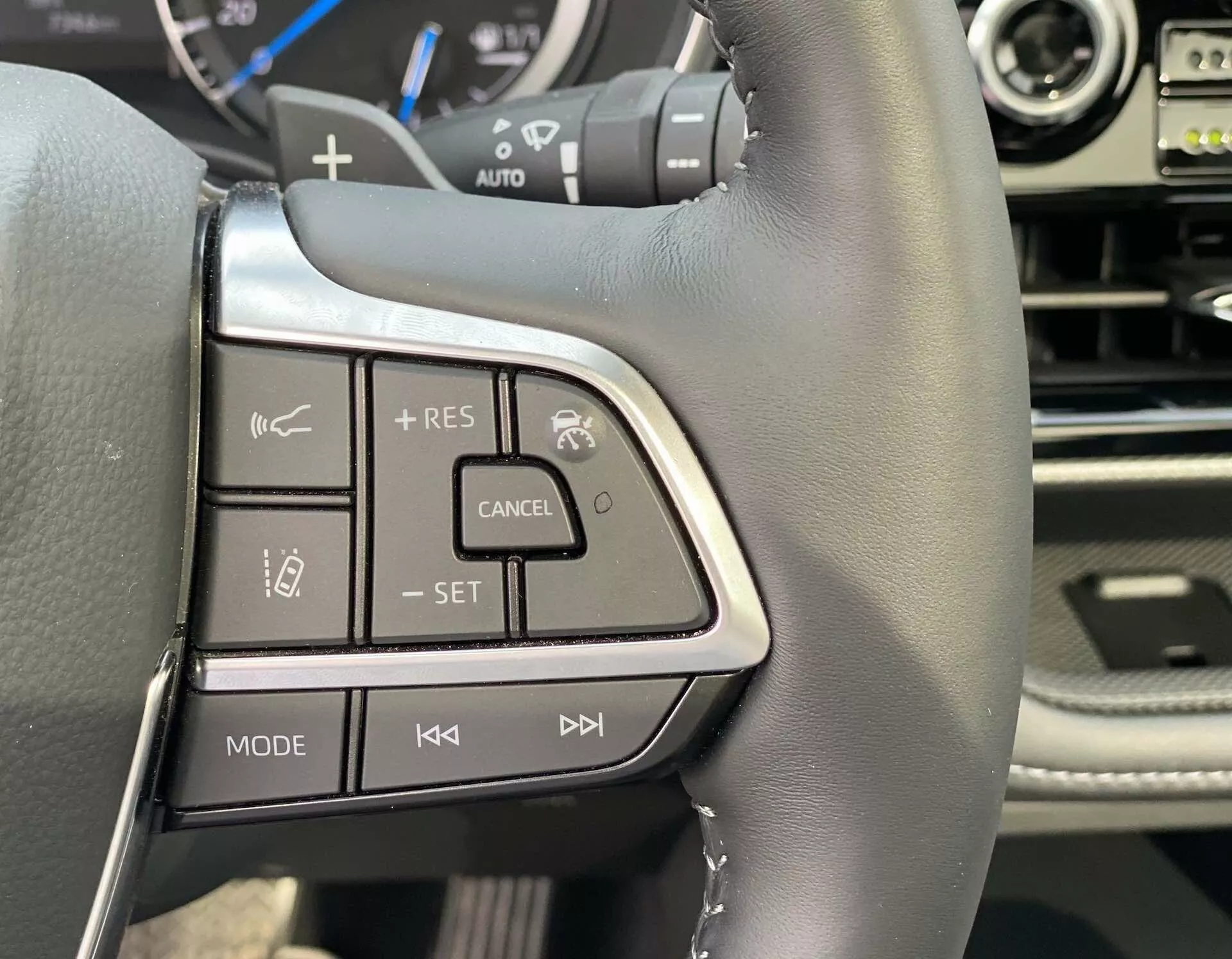 Zbliżenie lewej strony kierownicy w Toyocie Highlander 2021 2.5 Hybrid Business Edition, z czarną skórą z widocznymi szwami, srebrno/chromowymi akcentami, przyciskami adaptacyjnego tempomatu i systemu multimedialnego oraz częścią zestawu wskaźników i centralną deską rozdzielczą.