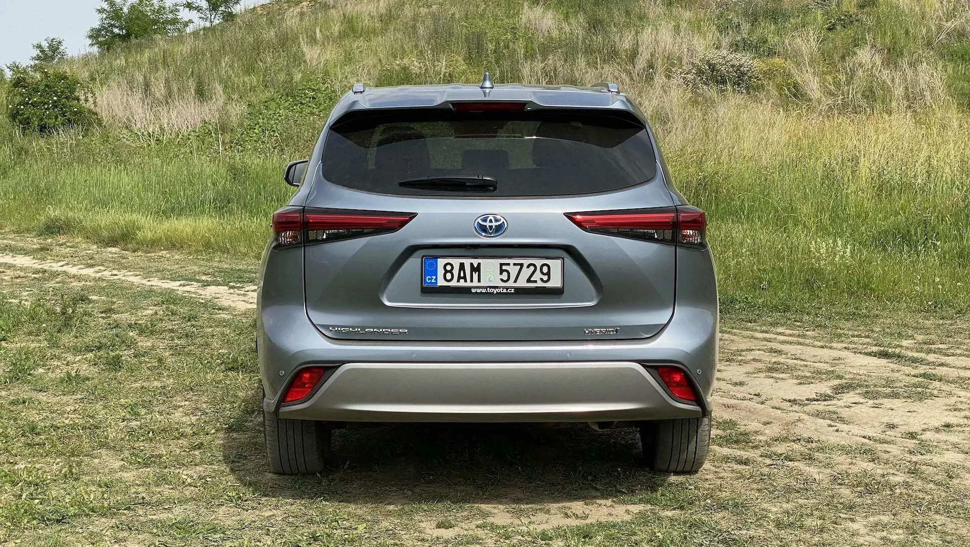 Widok tyłu srebrnego Toyota Highlander 2021 2.5 Hybrid Business Edition zaparkowanego na trawiastym terenie z zakurzoną ścieżką na pierwszym planie, uchwyconego w świetle dziennym z naturalnym oświetleniem. Nowoczesny SUV charakteryzuje się eleganckimi tylnymi światłami otaczającymi boki, wyraźnym tylnym spojlerem, zintegrowanymi odblaskami na zderzaku, kamerą cofania nad tablicą rejestracyjną i srebrną osłoną pod zderzakiem, co sugeruje jego zdolności terenowe i technologiczne zaawansowanie w zakresie wspomagania parkowania i systemów cofania.