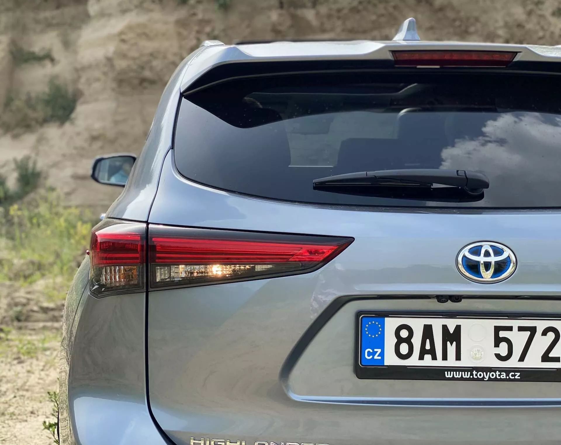 Zbliżenie tylnej sekcji Toyota Highlander 2021 Hybrid Business Edition w srebrnym kolorze, uchwycone pod kątem trzech czwartych, podkreślającym prawą stronę. Charakteryzuje się charakterystycznym tylnym światłem LED, tablicą rejestracyjną w stylu europejskim z Czech, oznaczeniem hybrydowym, systemem odmrażania tylnej szyby i wysoko umieszczoną lampą stopu na tle naturalnym terenie.