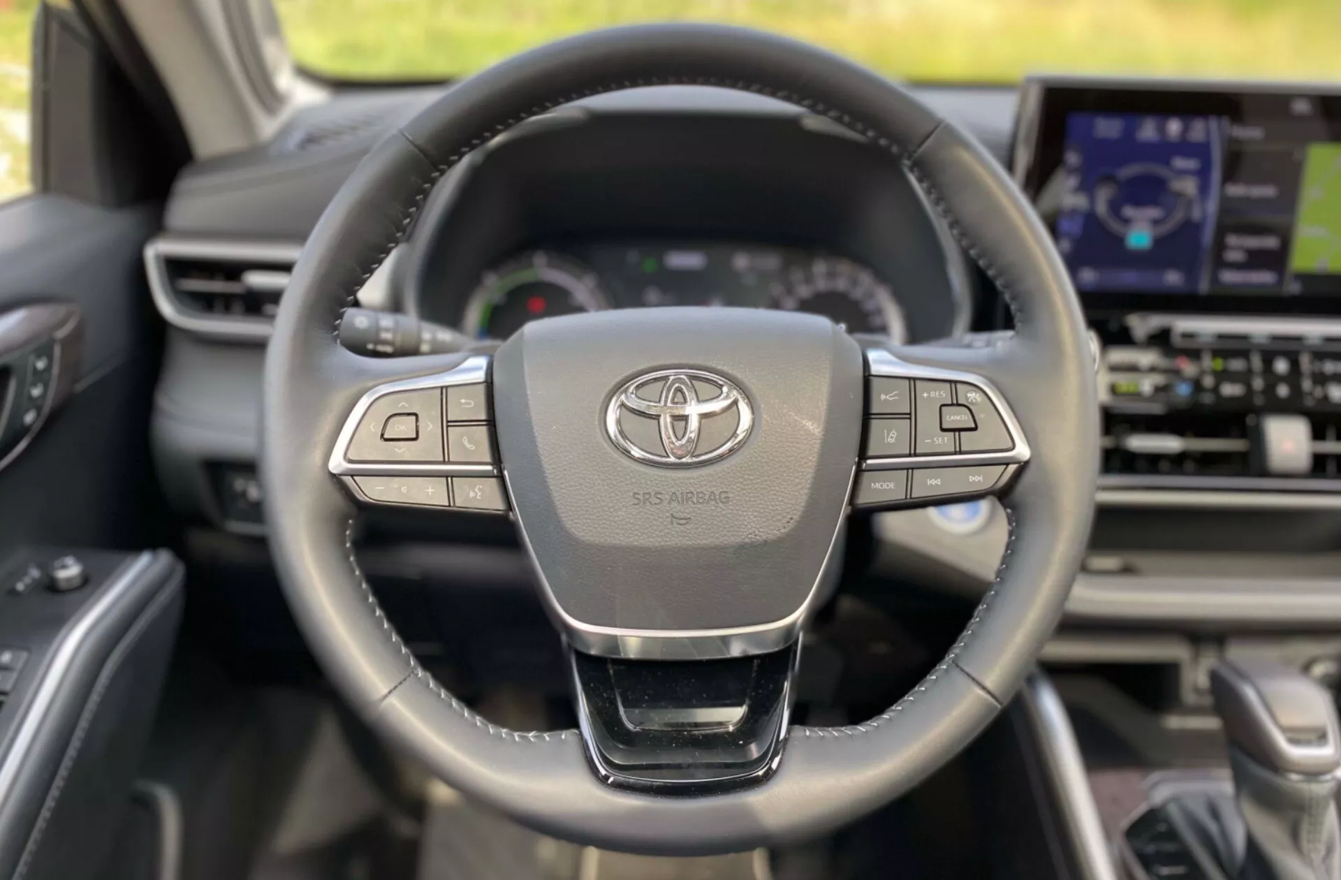 Wnętrze Toyoty Highlander 2021 Business Edition z perspektywy kierowcy, z nowoczesną, skórzaną trzyramienną kierownicą z wieloma przyciskami sterującymi, logo Toyota i napisem SRS AIRBAG. W tle deska rozdzielcza zawiera cyfrowy zestaw wskaźników i większy ekran informacyjno-rozrywkowy.