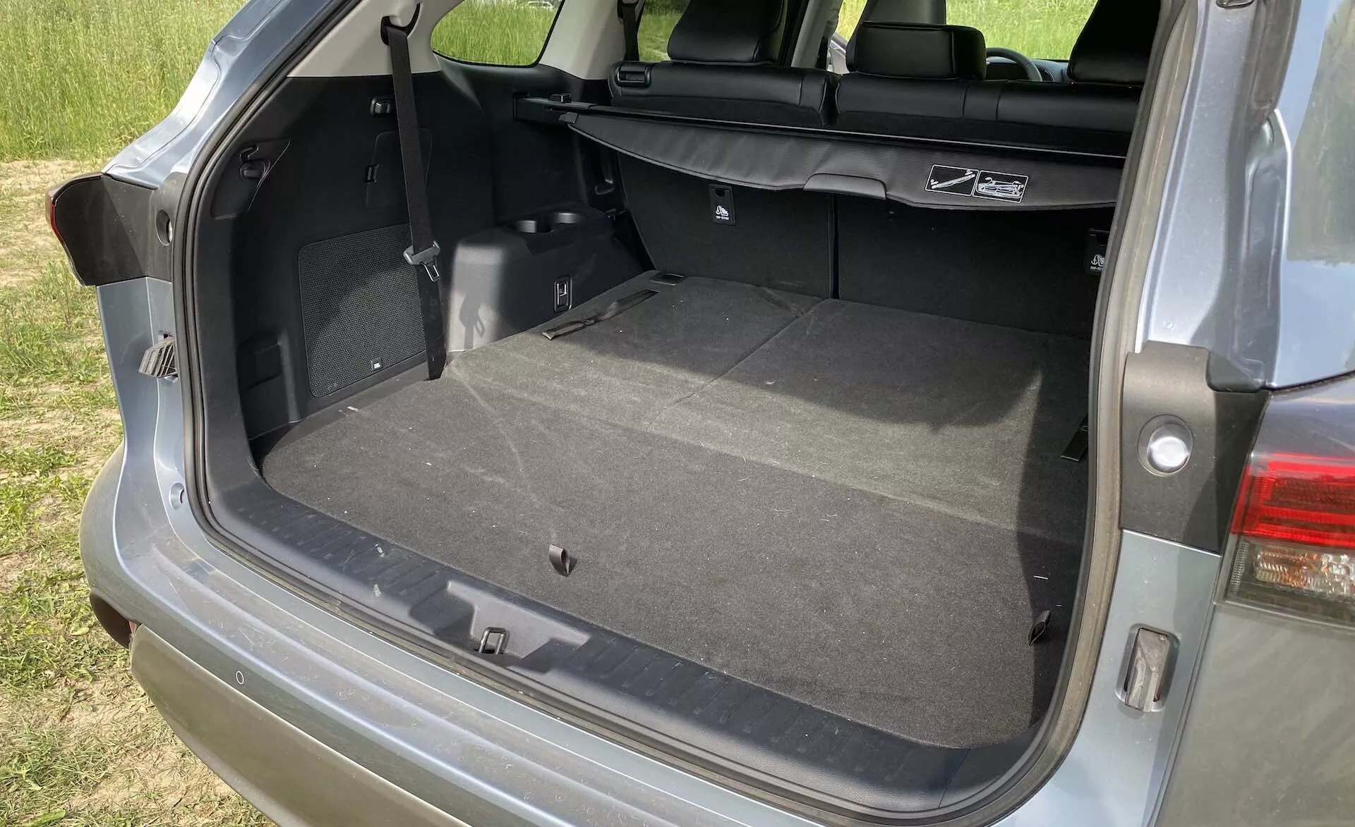 Zbliżenie na otwartą przestrzeń bagażową Toyota Highlander 2.5 Hybrid Business Edition 2021 z pionowymi tylnymi drzwiami i przestronnym tylnym przedziałem. Kąt z bocznej strony pasażera pokazuje czystą, wykładaną dywanami powierzchnię, z możliwie złożonymi tylnymi siedzeniami w celu powiększenia przestrzeni ładunkowej. Widoczne cechy to w tym wnętrzne światło, gniazdo 12V, uchwyt do podniesienia pokrywy schowka, zaczepy do fotelików dziecięcych i część nowoczesnego zestawu tylnych świateł LED.