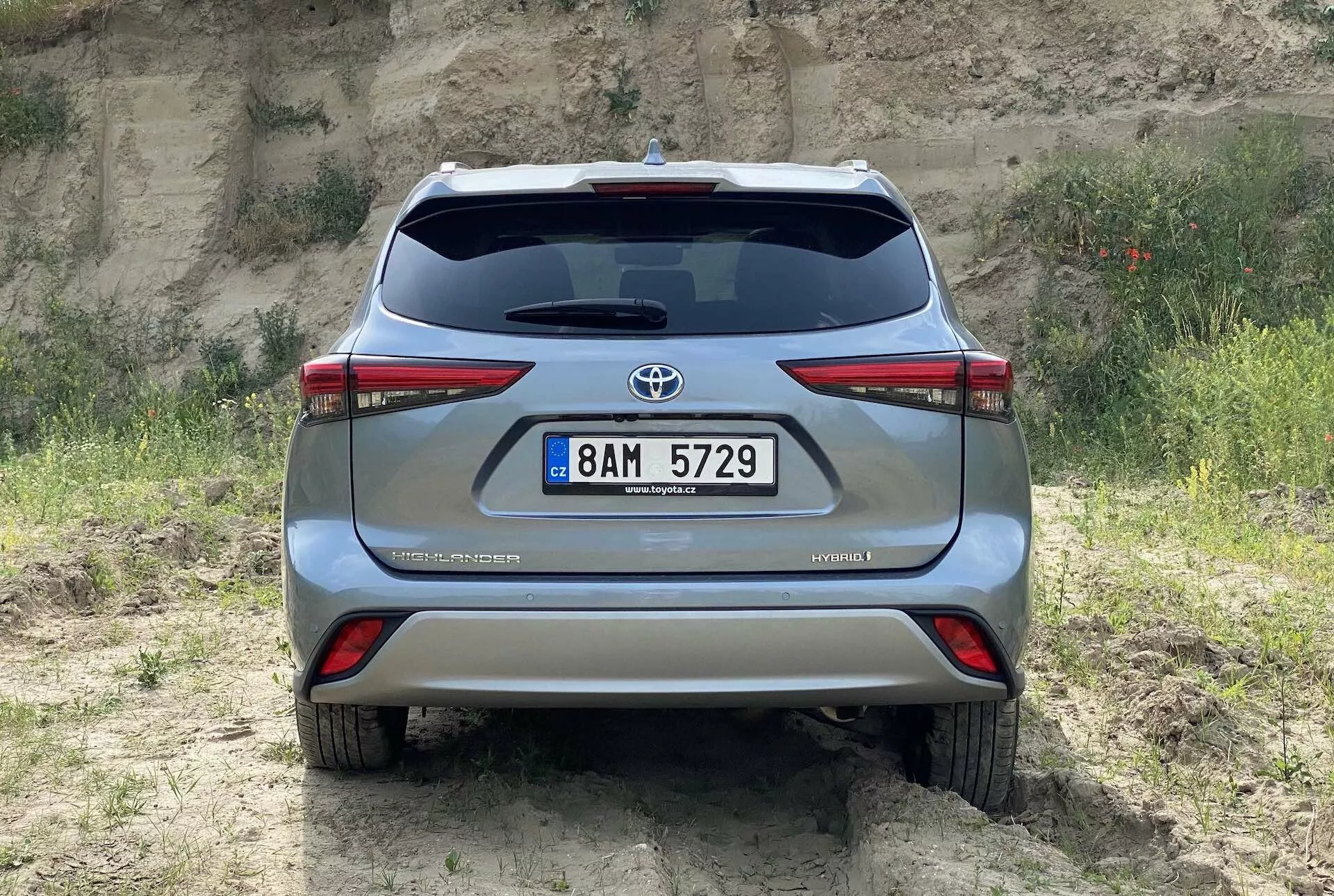 Tylny widok na srebrny Toyota Highlander 2021 2.5 Hybrid Business Edition uchwycony na wysokości oczu, pokazujący tylną klapę z emblematem Toyota, tylne światła, podwójny układ wydechowy, tablicę rejestracyjną na środku tylnego zderzaka oraz odznakę hybrydową, wszystko na naturalnym terenie z trawą i ziemią, podkreślając jego potencjał terenowy.