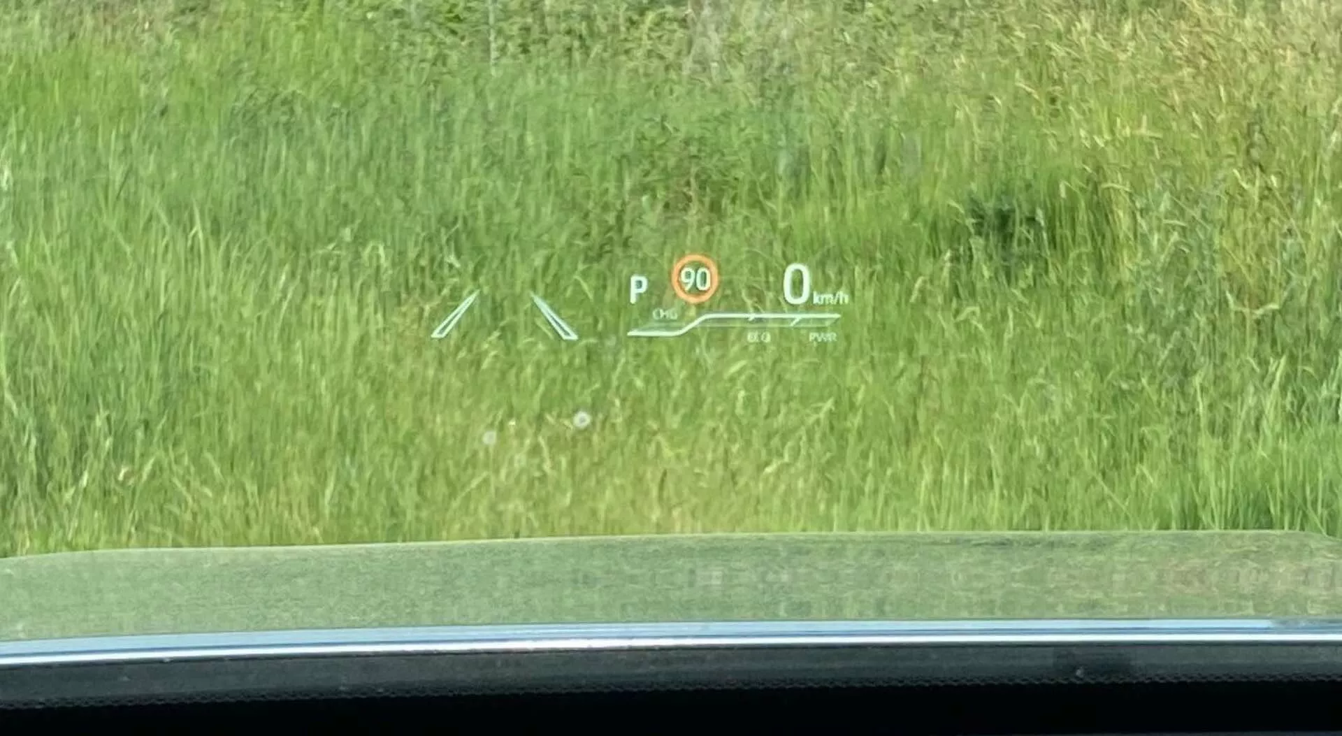 Widok wewnętrzny Toyoty Highlander 2021 2.5 Hybrid Business Edition skoncentrowany na wyświetlaczu head-up (HUD) wyświetlanym na przedniej szybie. HUD pokazuje podstawowe informacje dotyczące jazdy: wskaźnik 'P' oznaczający 'Park', znak ograniczenia prędkości '90', aktualną prędkość '0 km/h' oznaczającą, że samochód jest nieruchomy oraz symbole prawdopodobnie związane z systemem wspomagania parkowania lub asystentem pasa ruchu. Scena została uchwycona nieco na prawo od fotela kierowcy, z widokiem na trawiasty teren na zewnątrz.