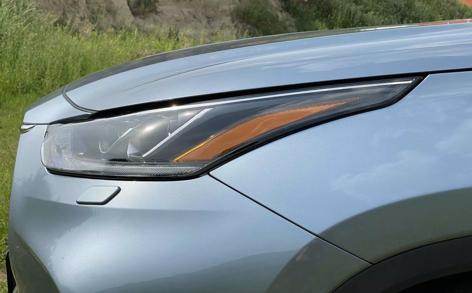 Zbliżenie przedniej lewej części Toyota Highlander 2.5 Hybrid Business Edition 2021, koncentrujące się na szczegółach zespołu reflektorów i części maski. Obraz jest wykonany z niskiego, lekko wznoszącego się i do przodu kąta, podkreślając nowoczesny reflektor LED lub laserowy z wieloma elementami oświetlenia, w tym bursztynowy sygnalizator skrętu. Maska ma metaliczny lakier z możliwościami perłowym wykończeniem i aerodynamicznymi liniami, podkreślając zaawansowaną technologię oświetlenia i luksusowy design.