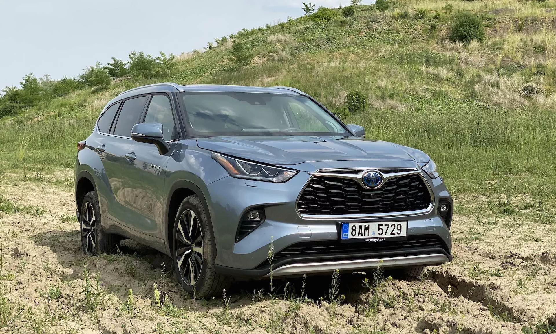 Trzy czwarte widoku z przodu szarego SUV-a Toyota Highlander 2.5 Hybrid Business Edition ustawionego na zewnątrz w terenie off-road z trawą i ziemią. Zdjęcie podkreśla wyraźny przedni grill z emblemą marki, charakterystyczne reflektory LED, które rozciągają się na boki, wieloszprychowe felgi aluminiowe o średnicy około 18-20 cali, rzeźbione linie nadwozia z konturowymi błotnikami, podwyższoną wysokość sugerującą możliwości napędu na wszystkie koła lub cztery koła, światła przeciwmgielne w przednim zderzaku, lusterka boczne z wbudowanymi kierunkowskazami i przyciemniane szyby na tylnych oknach. Zdjęcie zapewnia wyraźny widok na te cechy z poziomu oczu, z naturalnym oświetleniem pokazującym kolory i detale wzornicze samochodu.