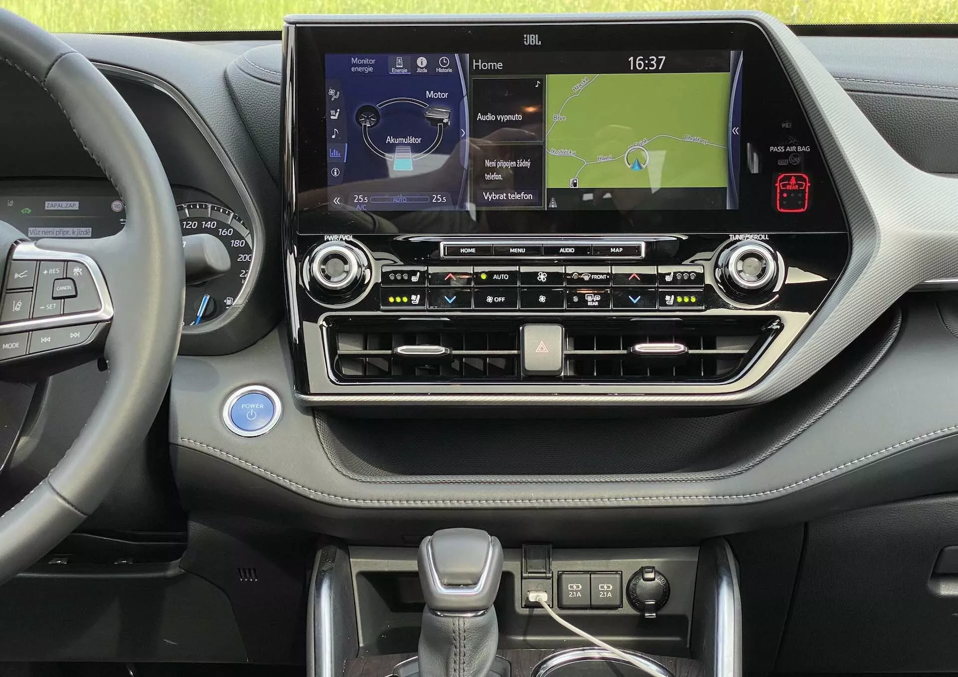 Wnętrze Toyoty Highlander 2021, skupiające się na konsoli centralnej i części strony kierowcy. Zdjęcie, zrobione z poziomu oczu, ukazuje krawędź kierownicy z wielofunkcyjnymi przyciskami, sugerującymi funkcje takie jak tempomat, sterowanie systemem audio i zarządzanie połączeniami bez użycia rąk. Konsola centralna wyposażona jest w duży ekran dotykowy z mapą nawigacyjną, wskazującą łączenie z mediami i telefonem. Poniżej ekranu dotykowego znajdują się przyciski i pokrętła do sterowania systemami klimatyzacji i audio, wraz z analogowym zegarem, metalowymi akcentami oraz deską rozdzielczą ze skóry lub miękkiego tworzywa. Dźwignia zmiany biegów jest nowoczesna i ergonomiczna, otoczona dodatkowymi przyciskami prawdopodobnie do trybów jazdy. Na dole można dostrzec uchwyt na napoje lub przestrzeń do przechowywania. Wnętrze jest czyste, dobrze oświetlone i zadbane.
